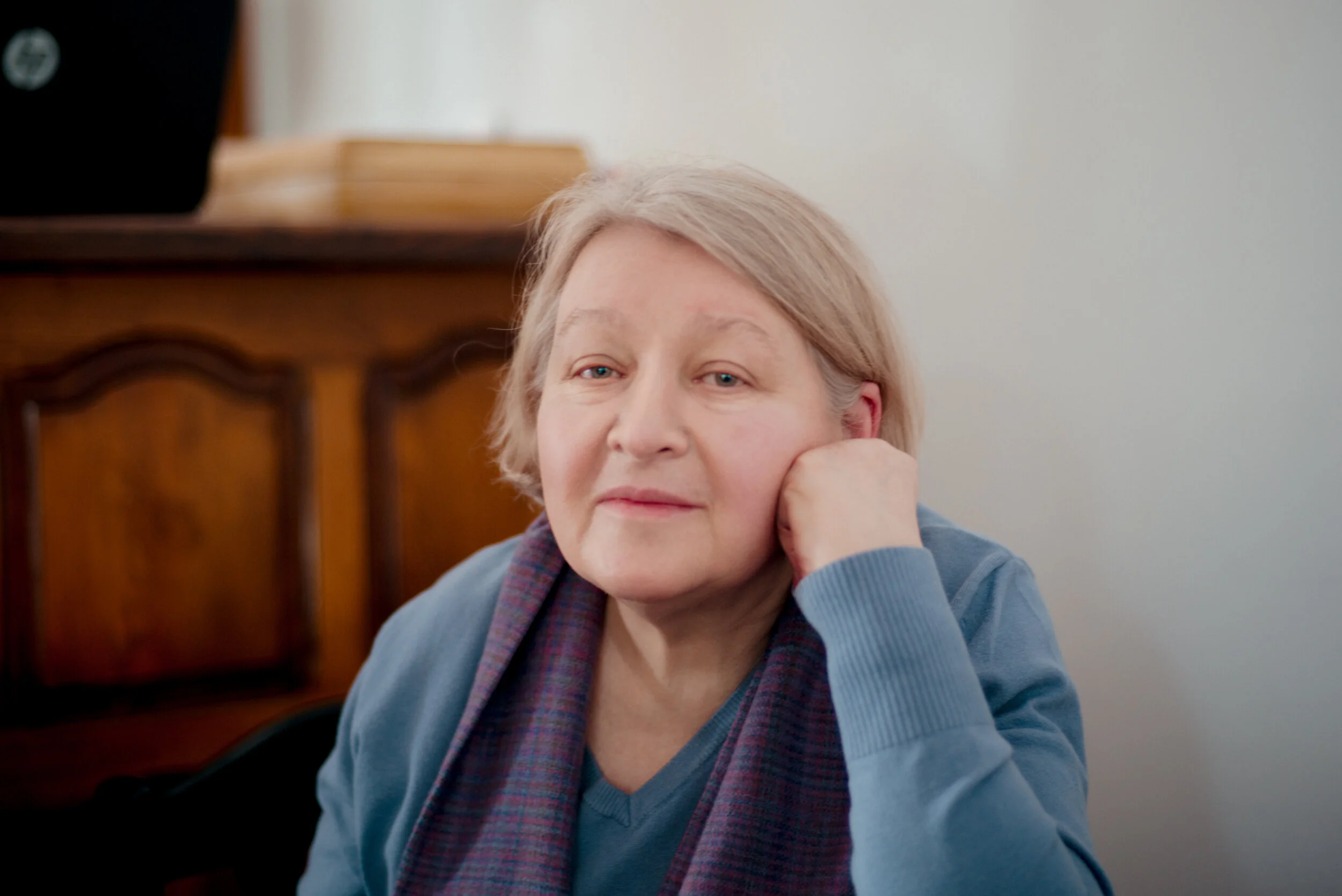 A middle-aged woman with short gray hair resting her chin on her hand, sitting indoors in front of a plain light-colored wall, with a wooden cabinet visible in the background.