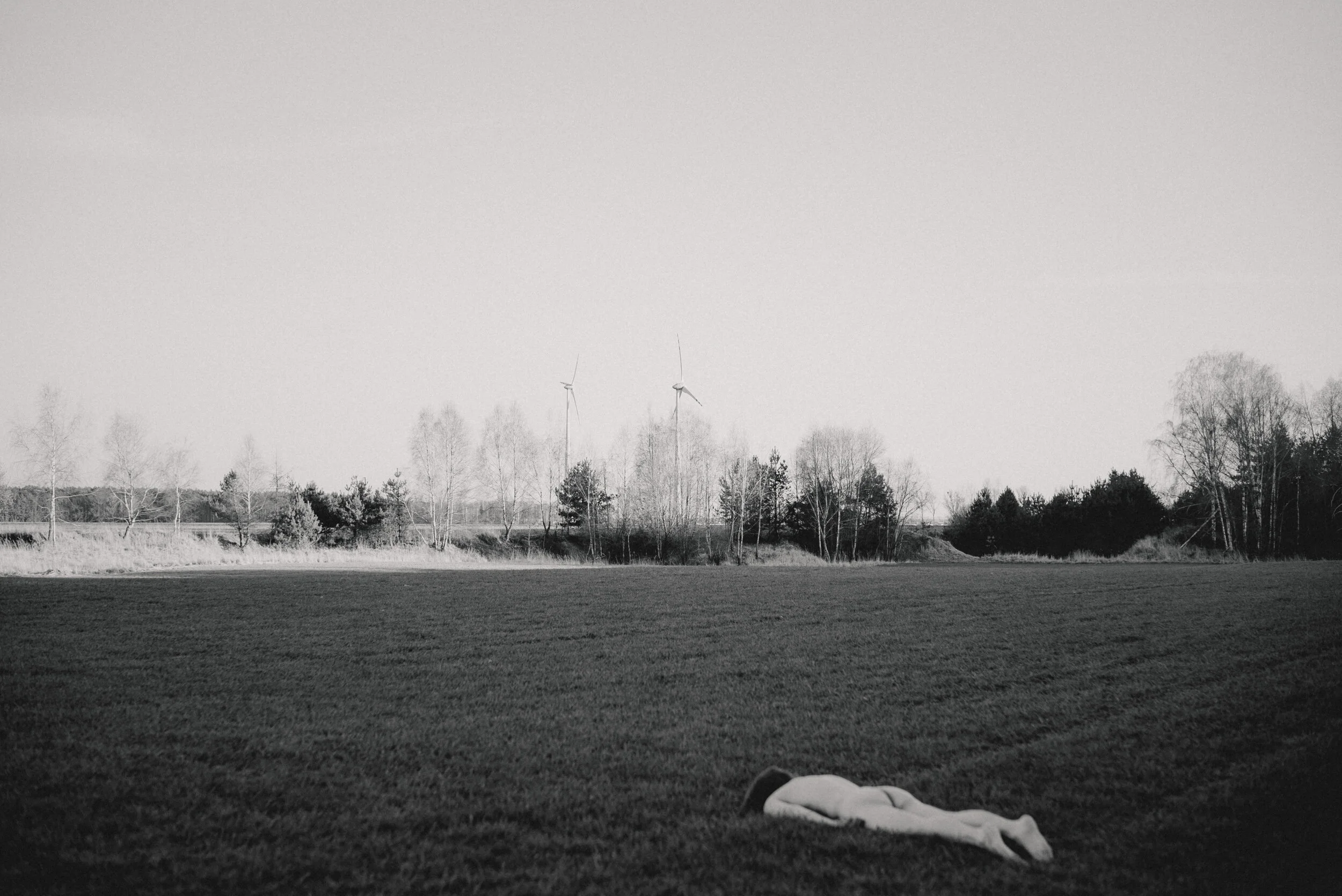 A black and white photograph of a field with a paper airplane on the ground in the foreground. In the background, there's a line of trees and wind turbines under a clear sky.