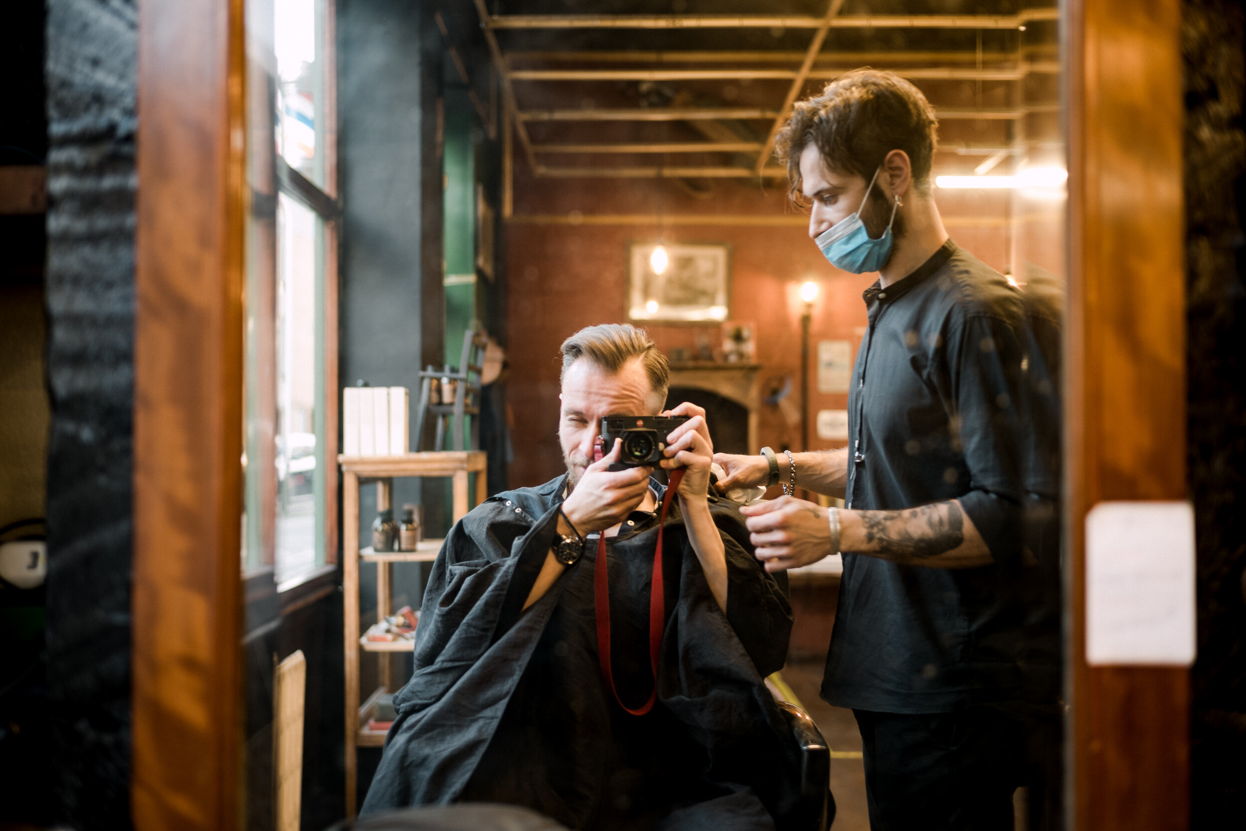 A man with a beard sitting in a barber's chair taking a selfie in a mirror, with a barber standing next to him wearing a face mask.