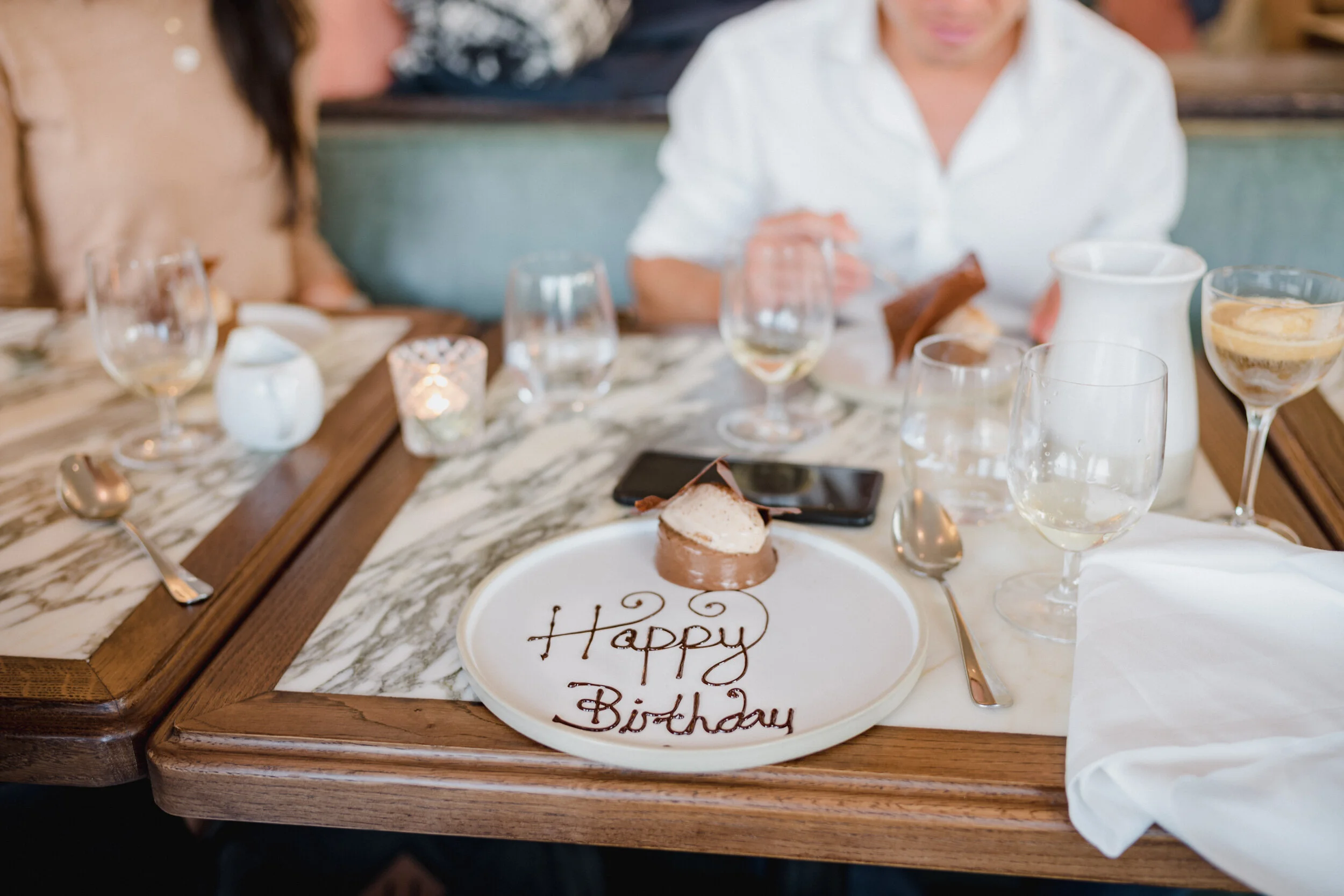 A birthday celebration at a restaurant with a table set with glasses, a dessert plate with a small cake, and a message that reads 'Happy Birthday' on a plate. Two people are partially visible, and the focus is on the table setting and the dessert.