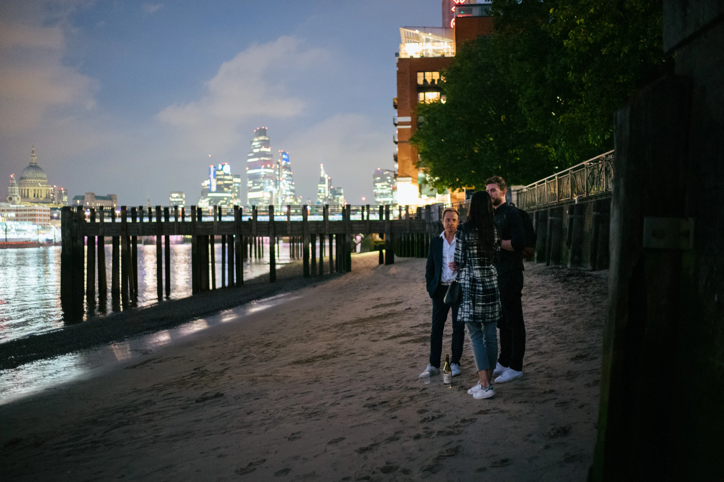 Three people stand and converse on a sandy beach at night, with a cityscape illuminated in the background, and a city building with lights to the right.