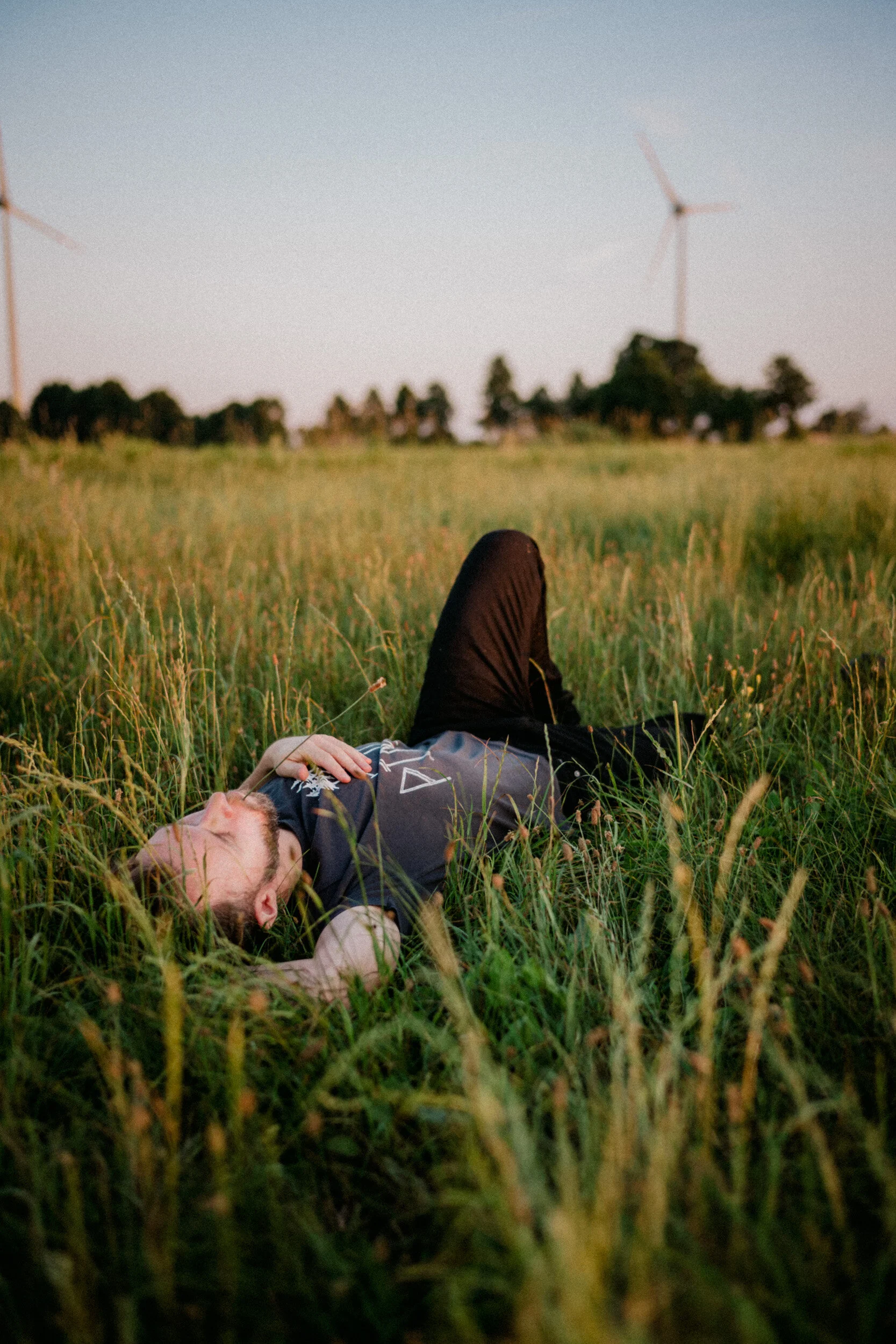 Person lying in a grassy field with wind turbines in the background, during sunset or sunrise.
