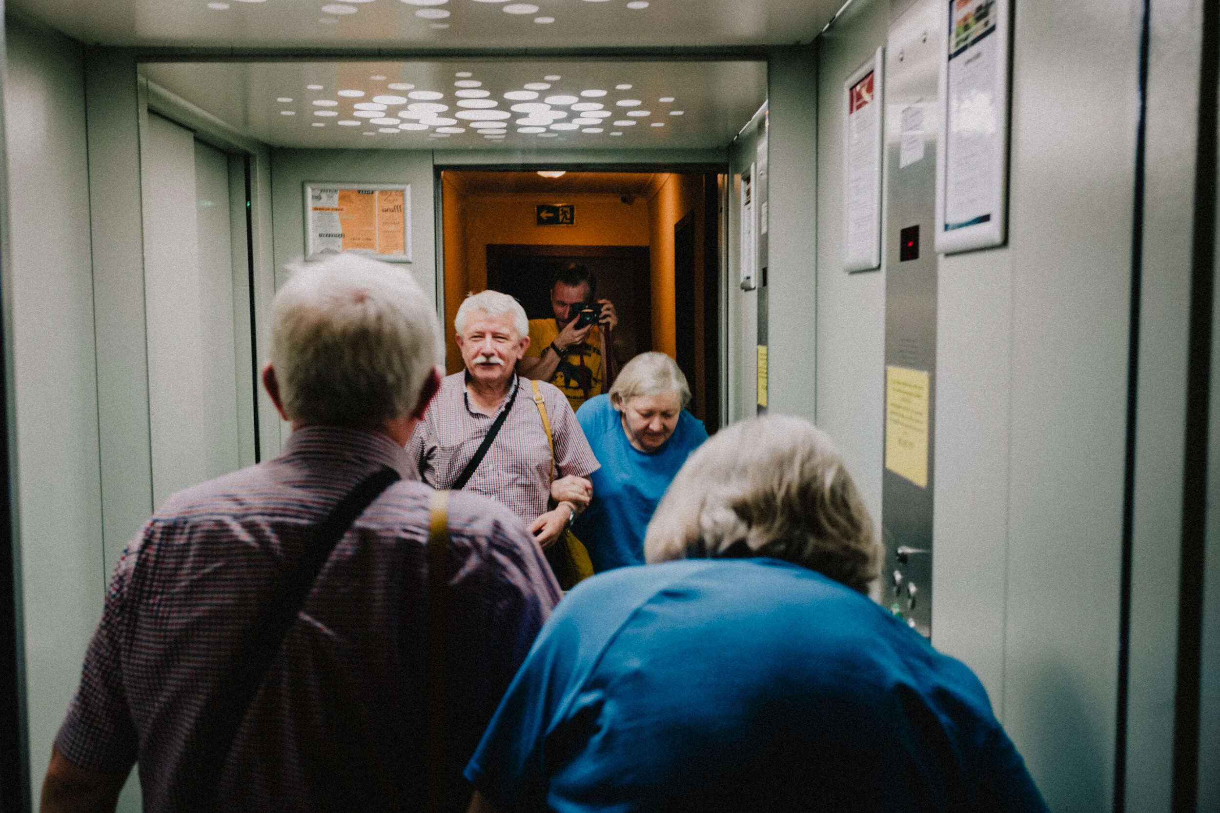 Elderly people boarding an elevator, with an elderly man and woman in the foreground and a man taking a photo in the background.