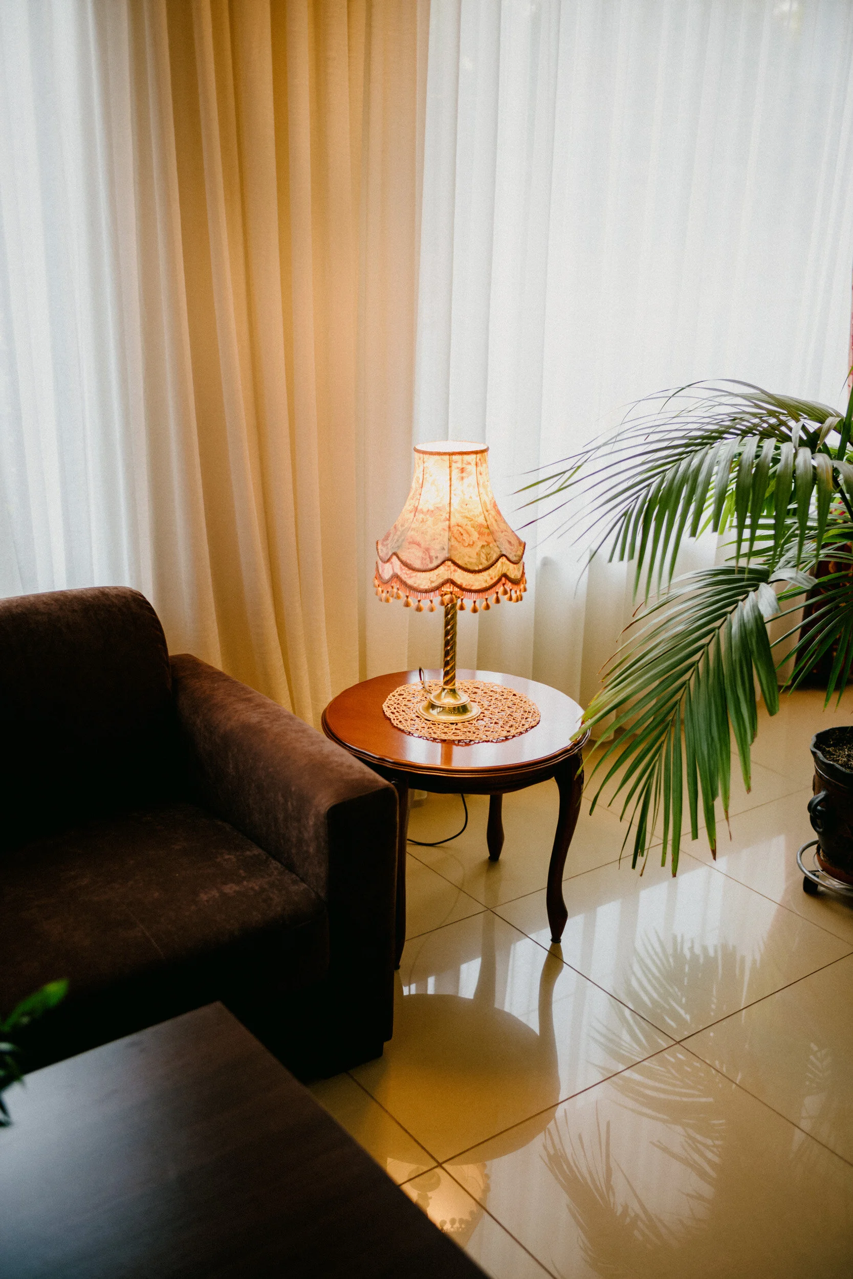 A cozy living room corner with cream curtains, a vintage table lamp on a wooden side table with a crocheted doily, and a green potted palm plant. Part of a brown sofa is visible in the foreground and there is reflected light on the tiled floor.