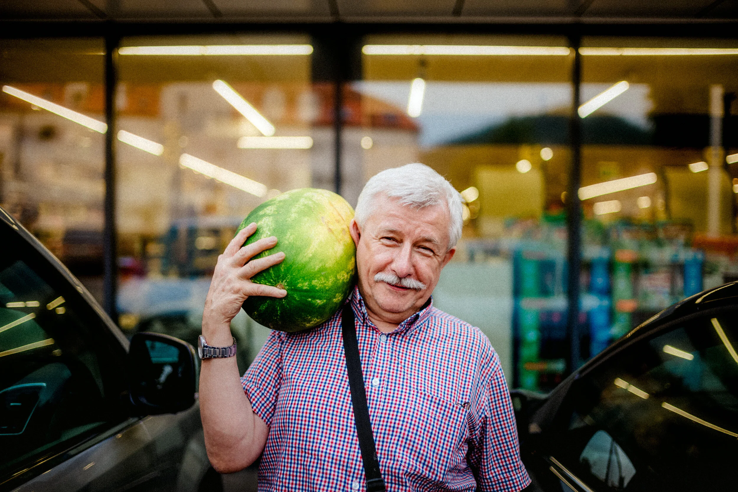 An elderly man with white hair and a mustache, smiling, carrying a large green watermelon on his shoulder, standing inside a grocery store near a black vehicle.