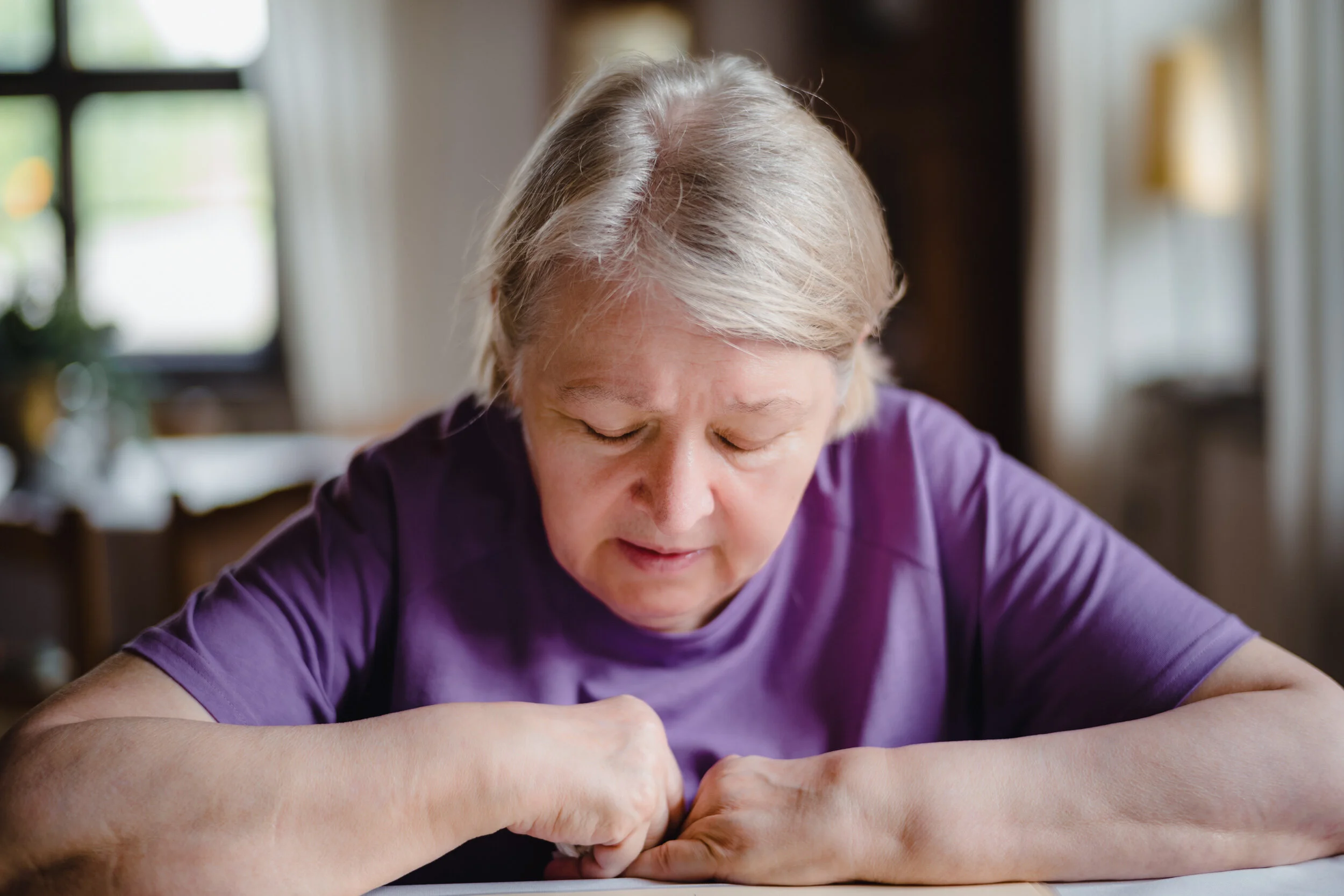 An elderly woman with gray hair, wearing a purple shirt, sitting at a table, focused on an activity with her hands, in a room with a blurred window and yellow curtain in the background.