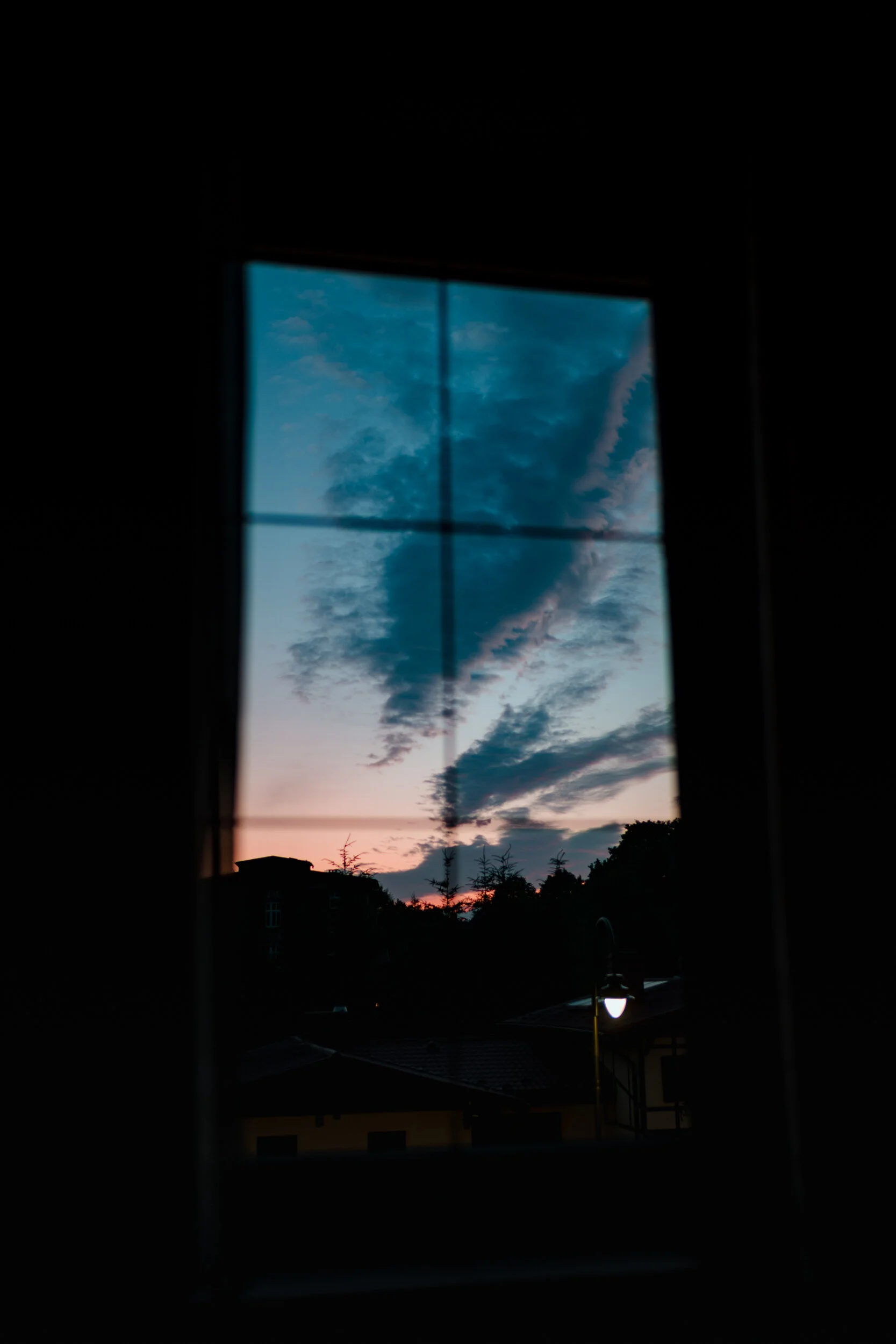 Looking through a window at a colorful sky during sunset, with dark silhouettes of trees, rooftops, and a street lamp outside.