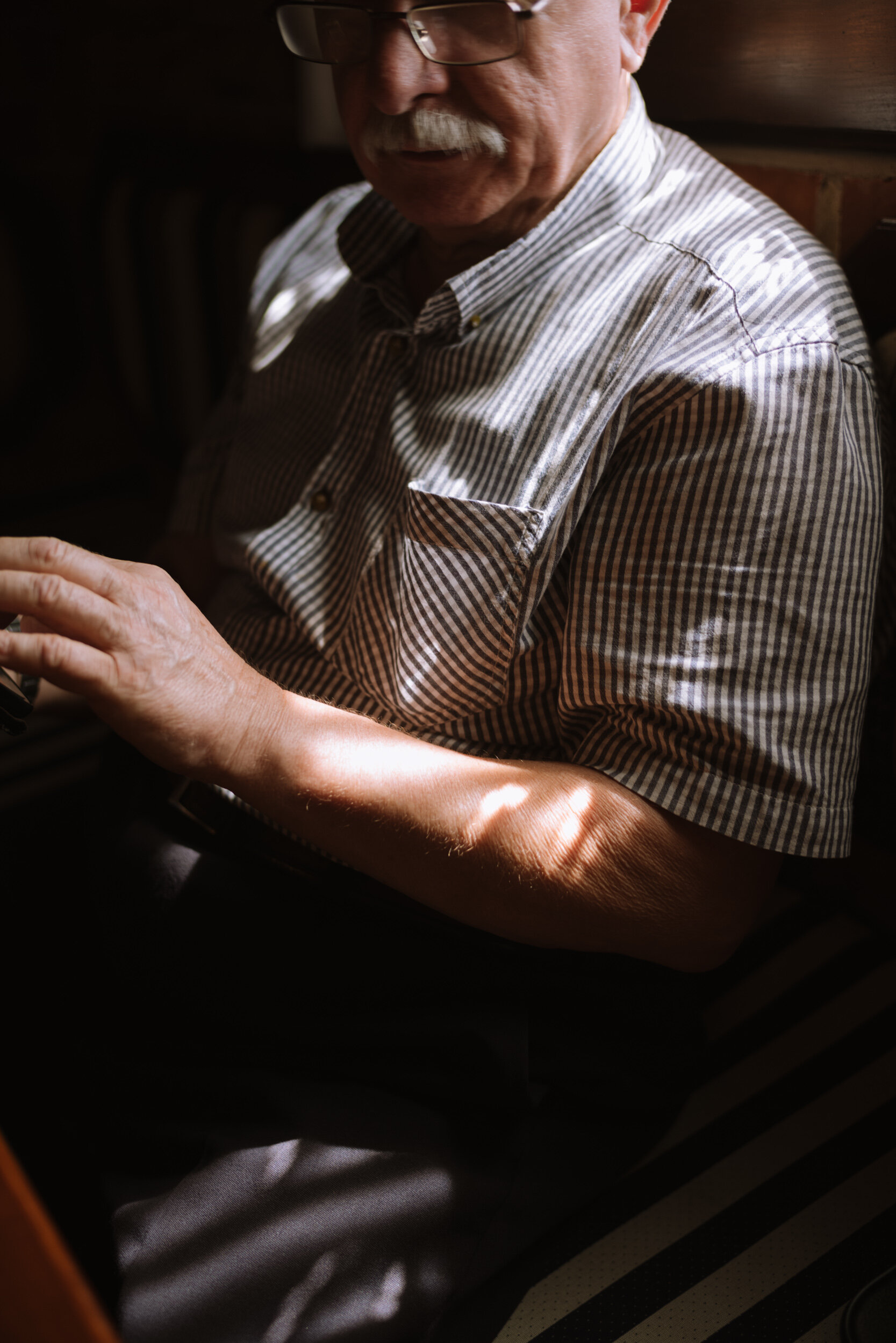 Close-up of an elderly man with glasses, a mustache, and striped shirt, sitting in a dimly lit room, looking down at something in his hand.