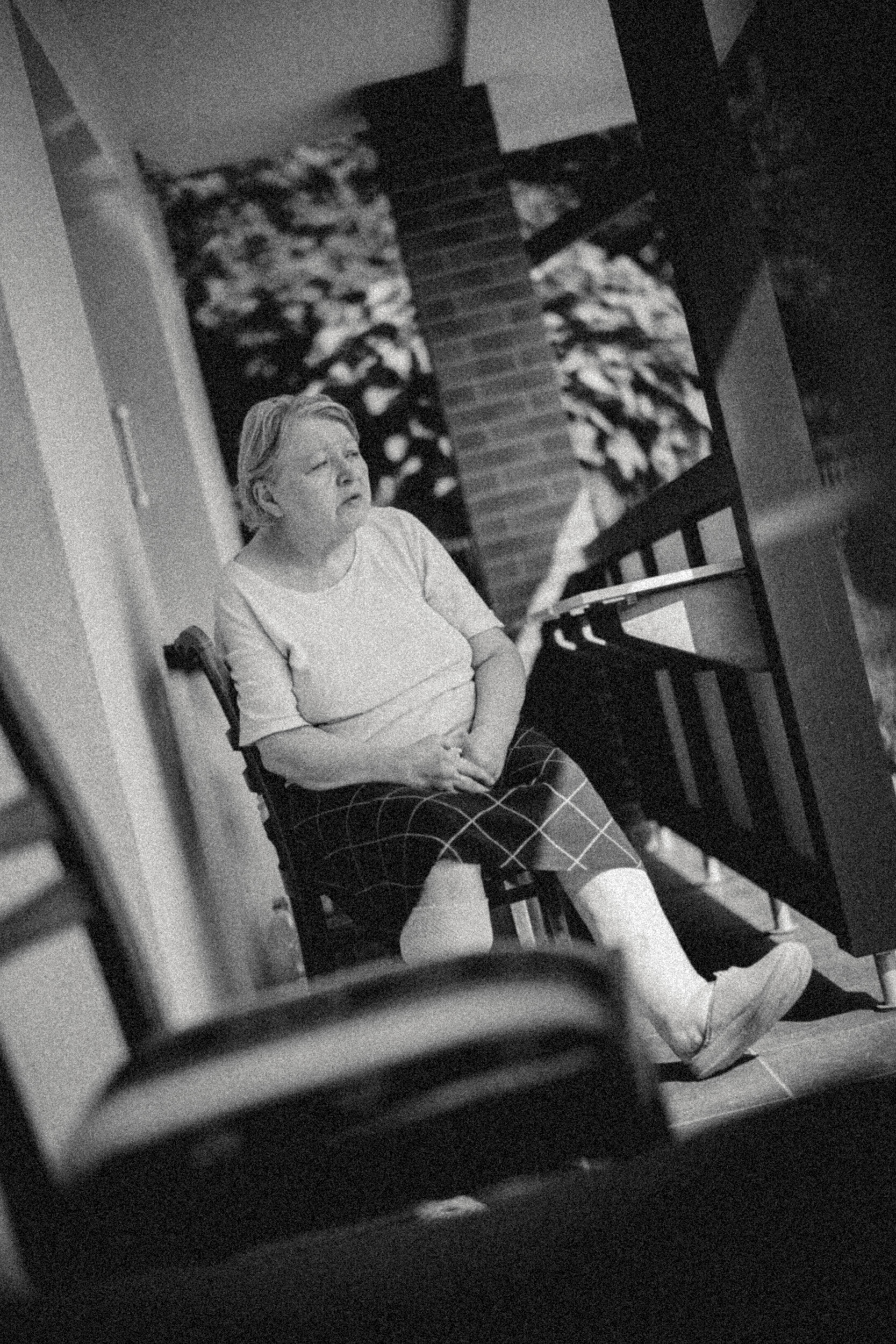 A woman with short hair wearing a light-colored shirt and checkered socks sitting on the floor with her arm resting on her lap, seen through a mirror with a warped reflection, in a dimly lit room with a staircase in the background.