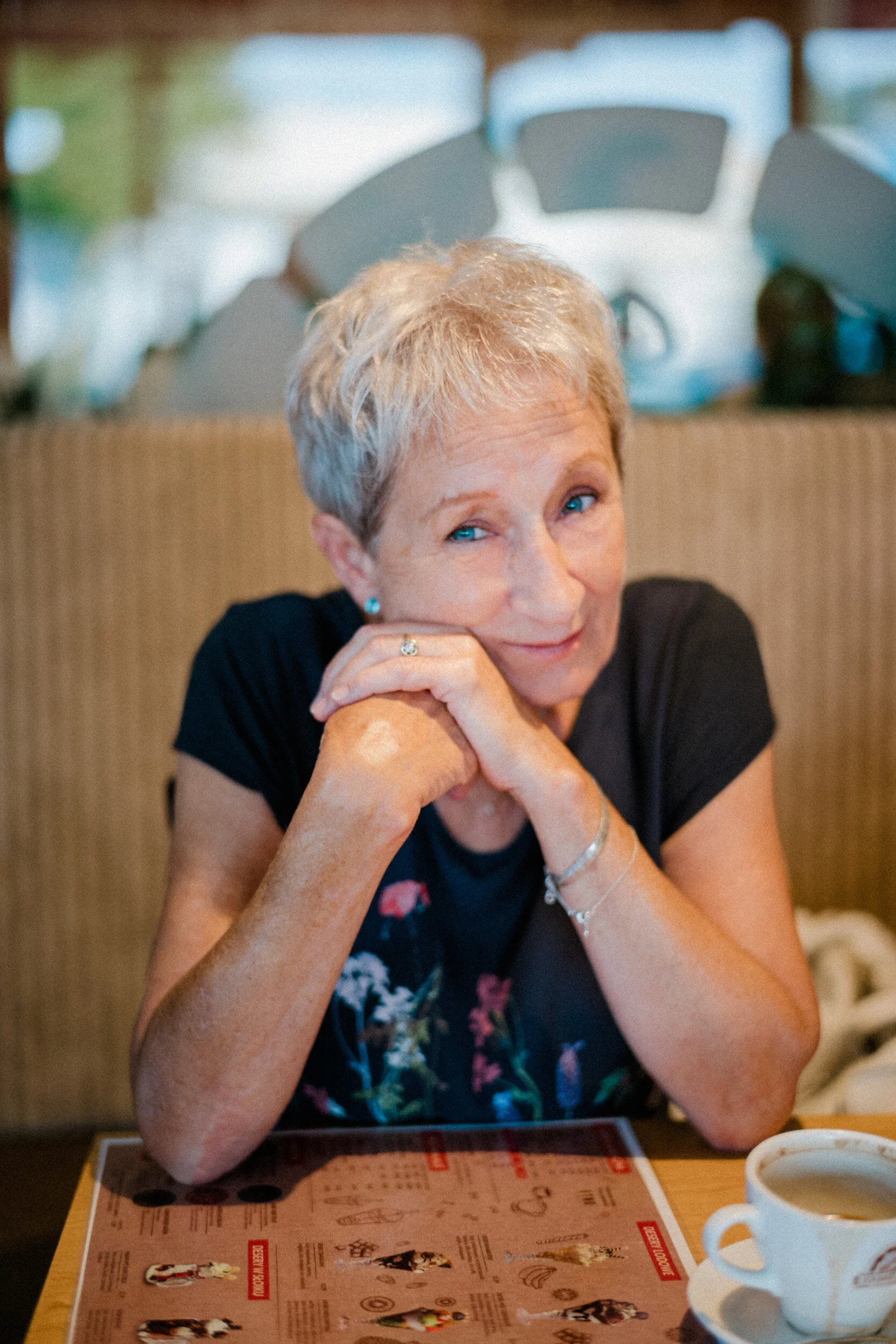 An elderly woman with short gray hair and blue eyes sitting at a table in a restaurant, resting her chin on her hands, with a menu in front of her and a coffee cup nearby.
