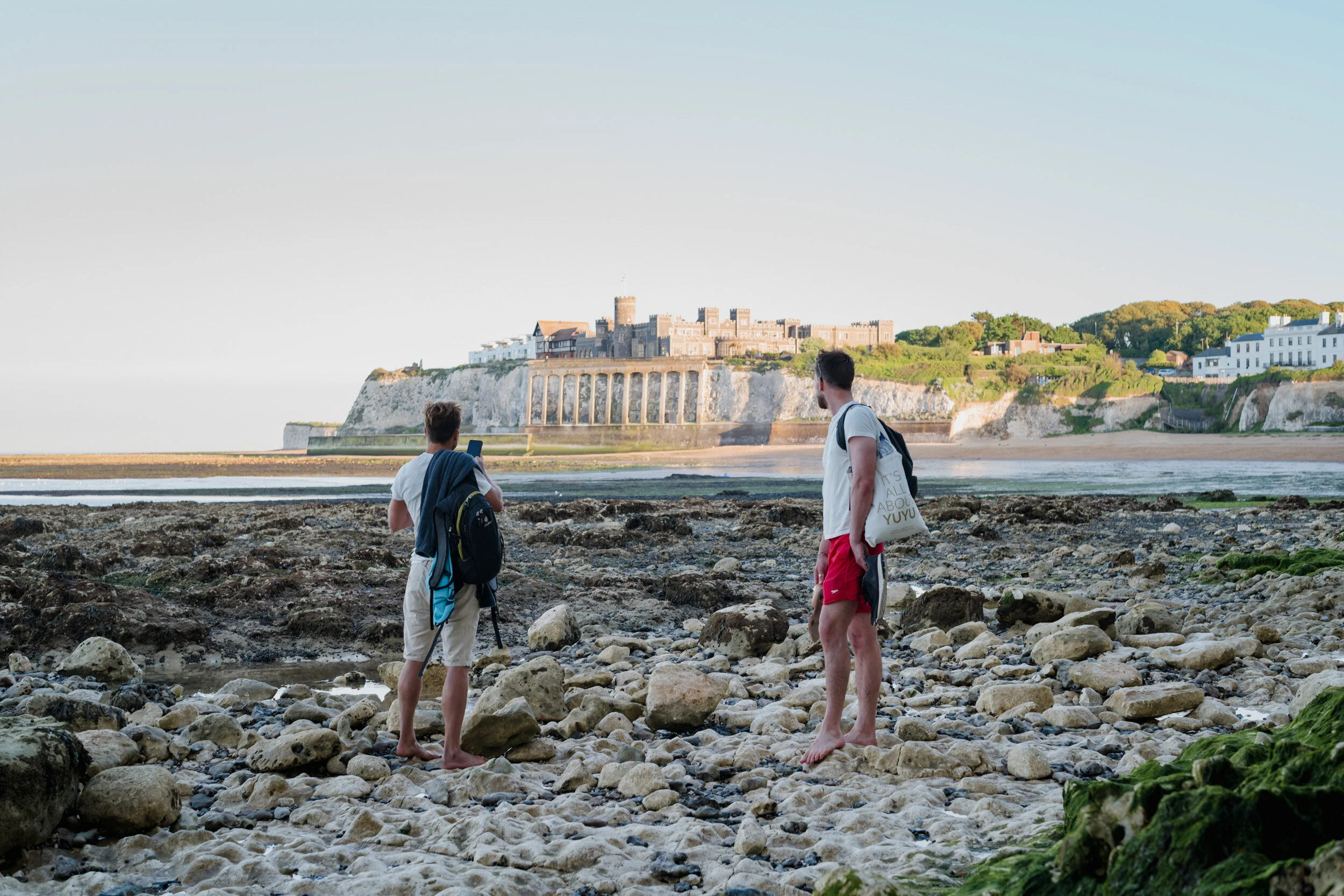 Two young men standing on a rocky beach near the tide line, looking toward a castle on cliffs in the distance, with one of them taking a photo with his smartphone.