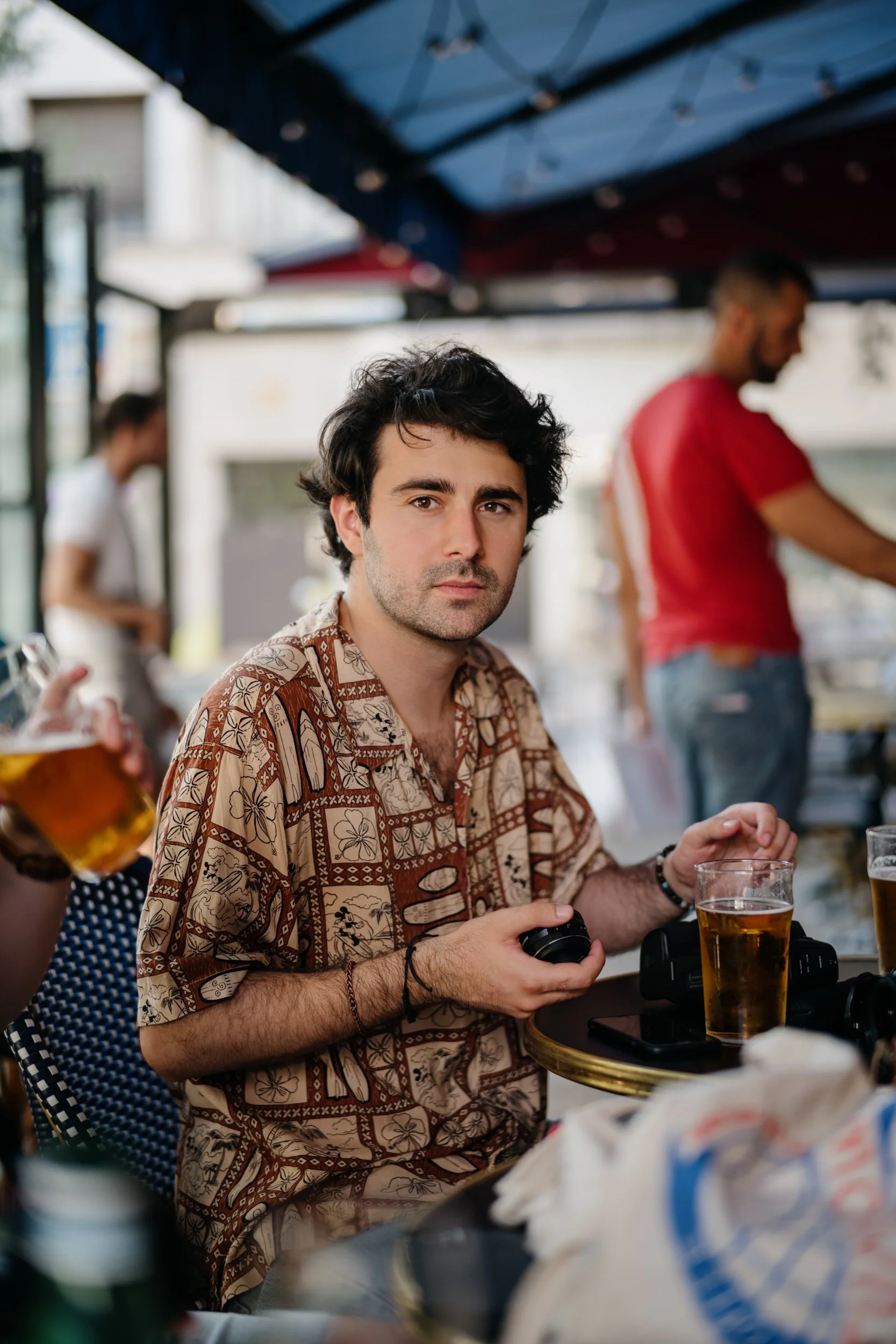 A man with dark curly hair and light skin, wearing a patterned shirt, sitting at an outdoor table with a drink in front of him, holding a camera in his hands. There are blurred people and objects in the background, including a person with a beard and