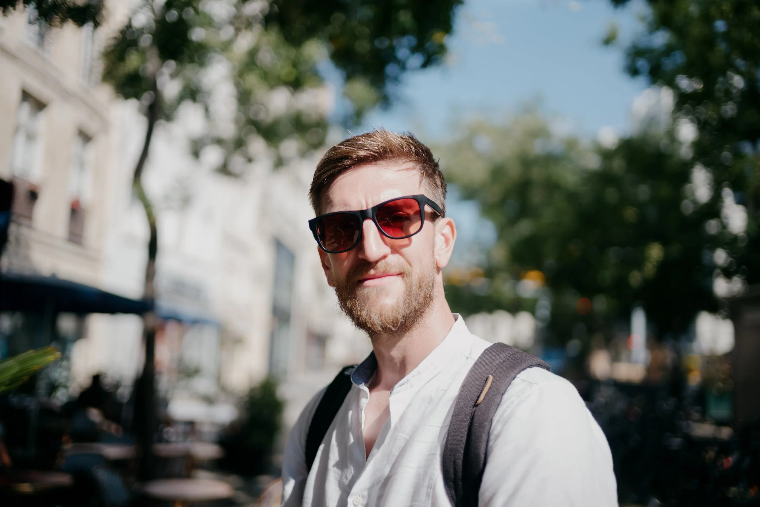 Young man with brown hair, beard, wearing sunglasses, a white shirt, and a backpack, outdoors on a sunny day, with trees and buildings in the background.