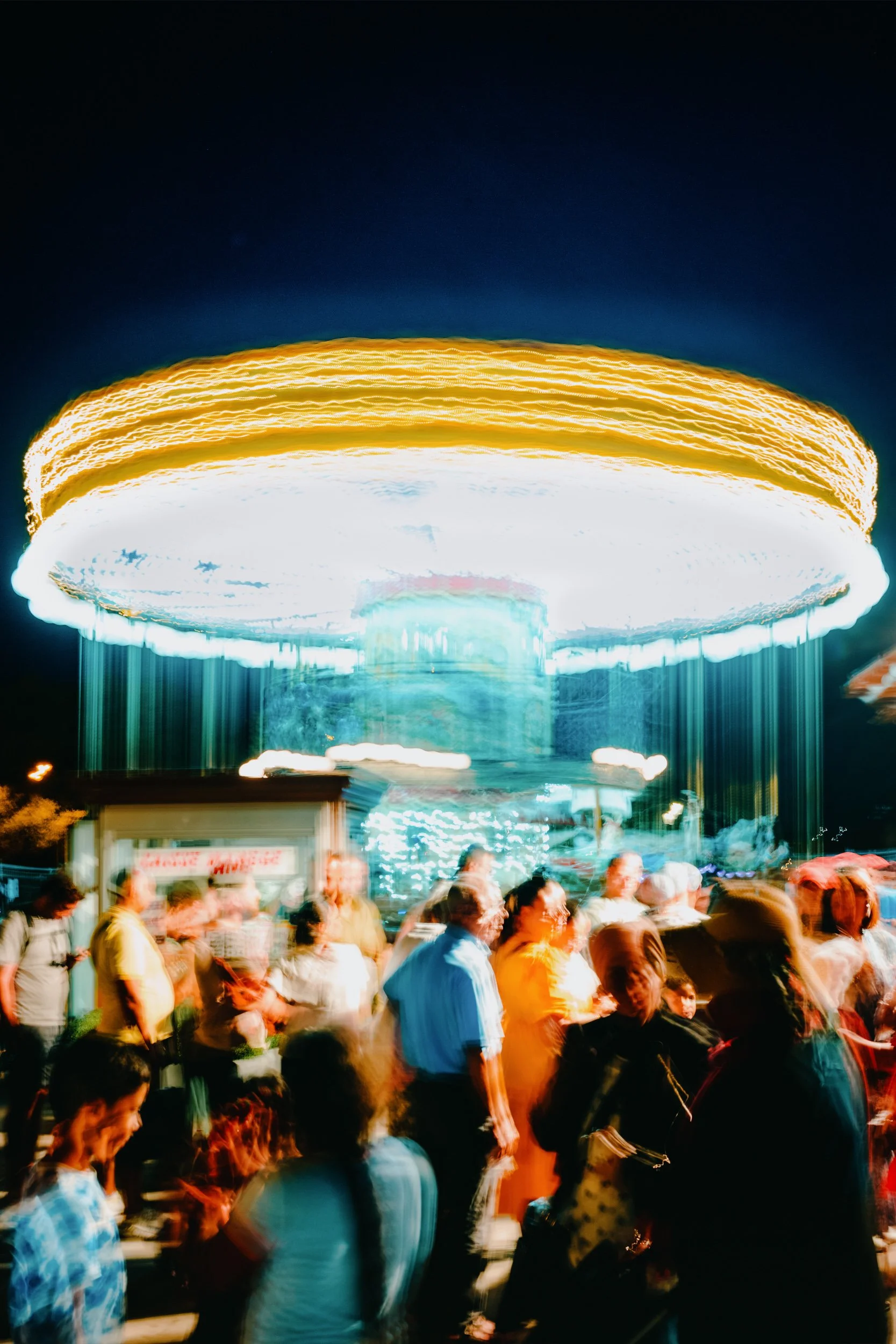 A brightly lit amusement park ride spinning at night with people in line and a dark sky in the background.