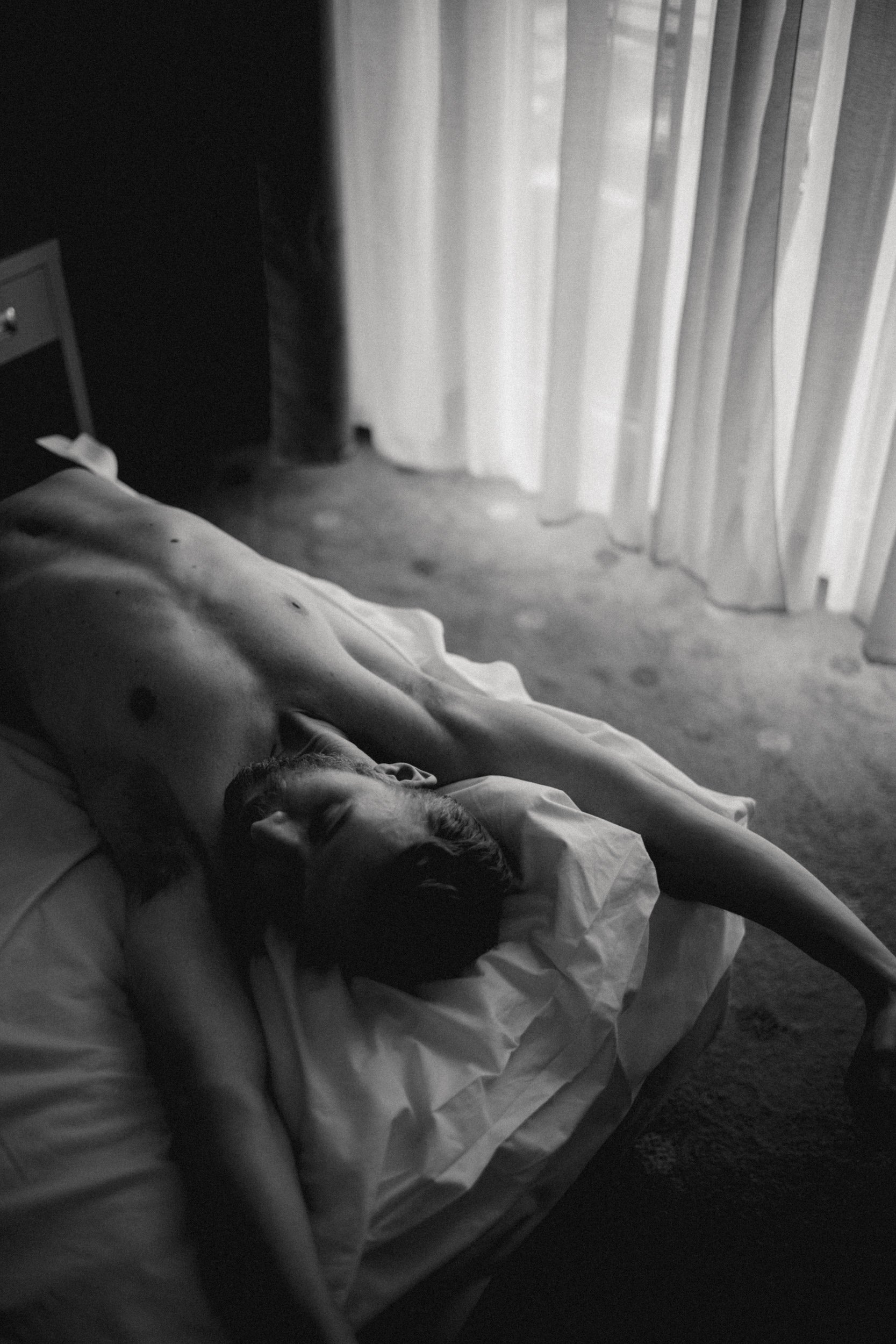 A person lying on a bed with their arm stretched out. The room has a window with vertical blinds. The photo is in black and white.
