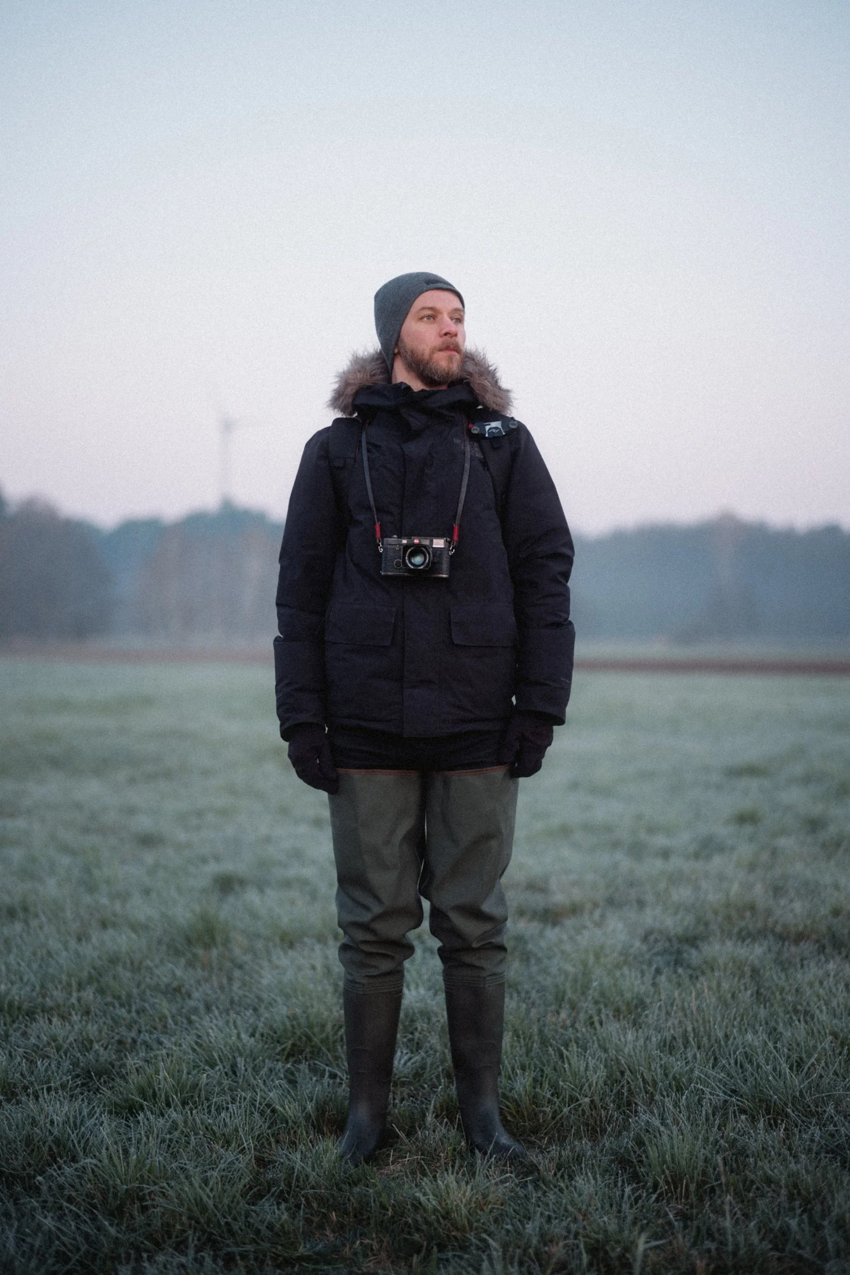 A person standing outdoors in a grassy field, wearing a dark jacket, gloves, rubber boots, and a gray beanie, with a camera hanging around their neck. The background is misty and slightly obscured with trees and a cloudy sky.