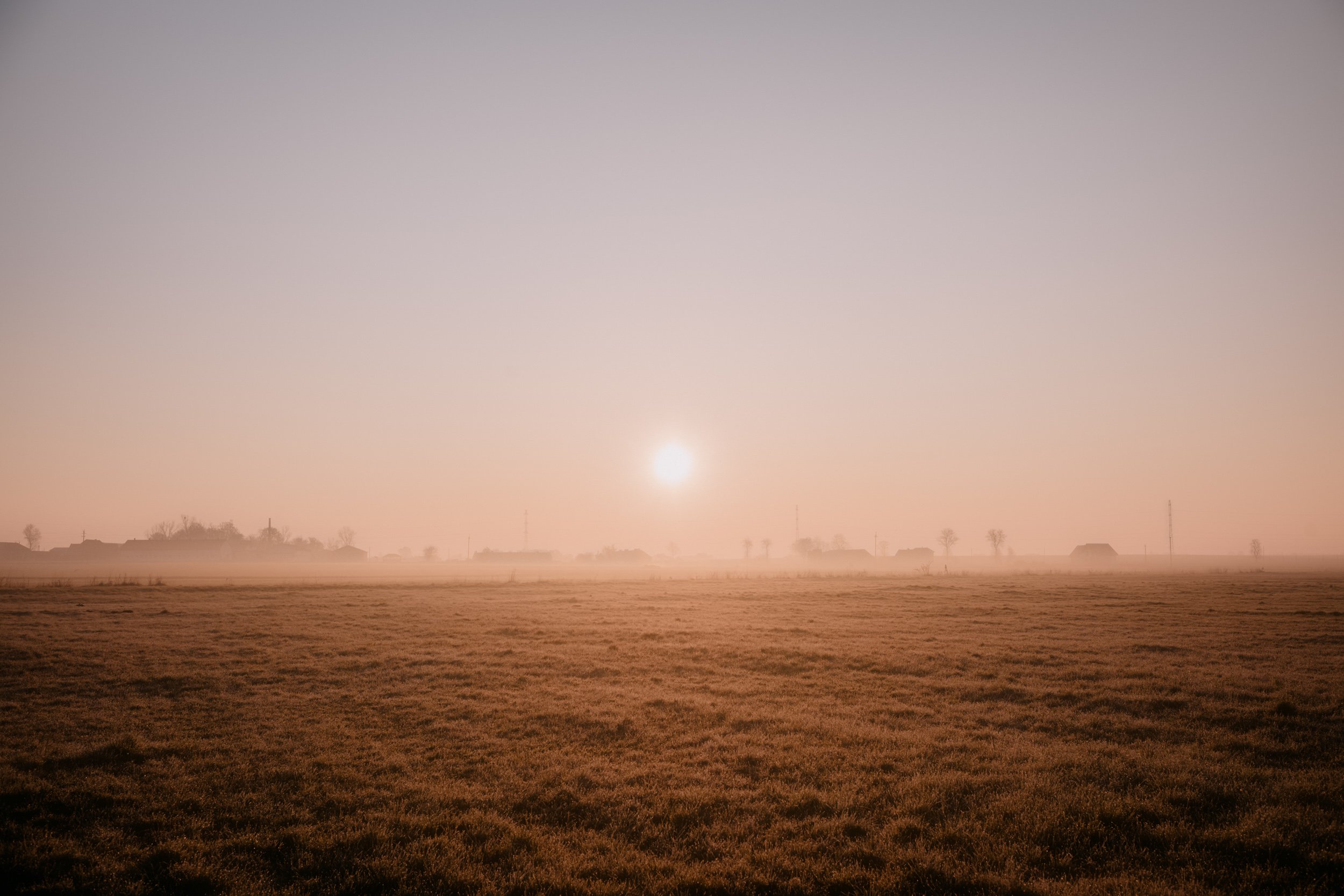 A wide view of a field during sunrise or sunset with a misty atmosphere, houses in the distance, and a clear sky.