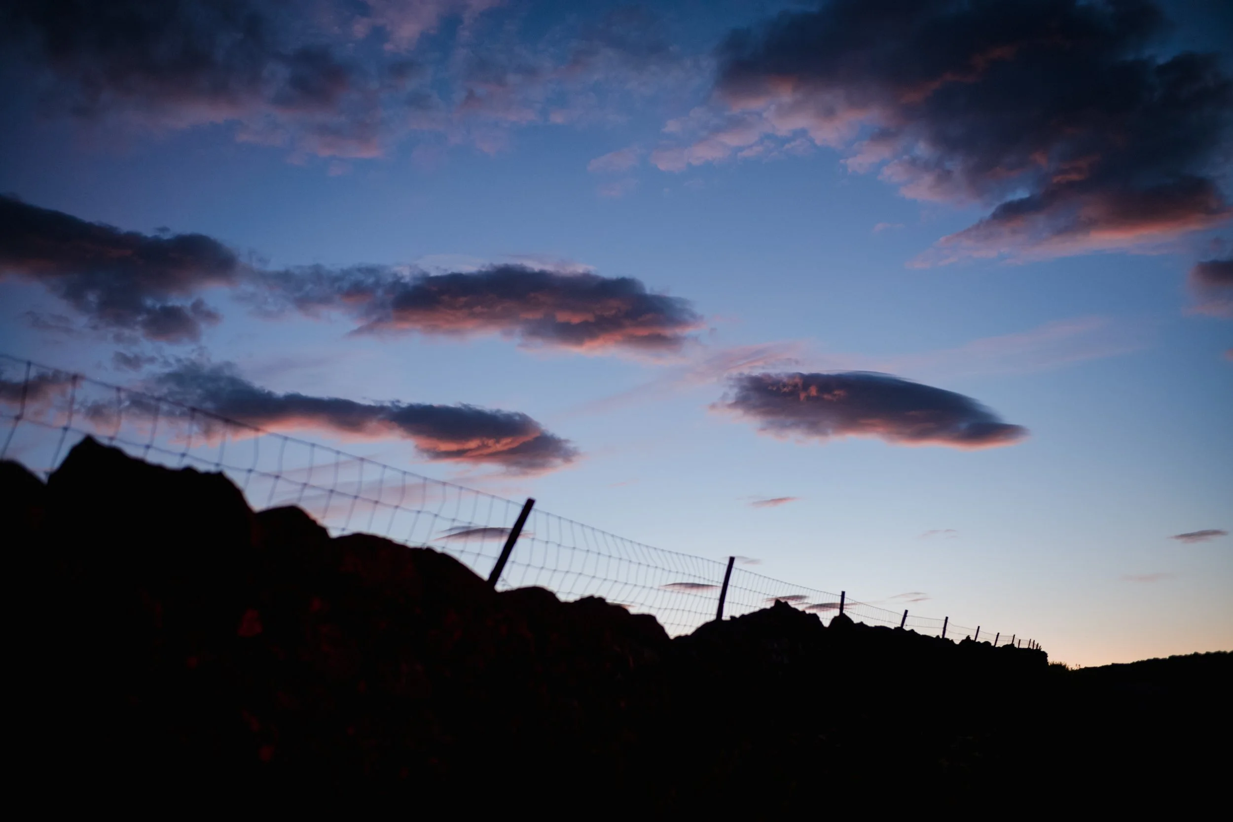 Silhouette of a hill with a fence, darkening sky at sunset with scattered dark clouds and pinkish hues.