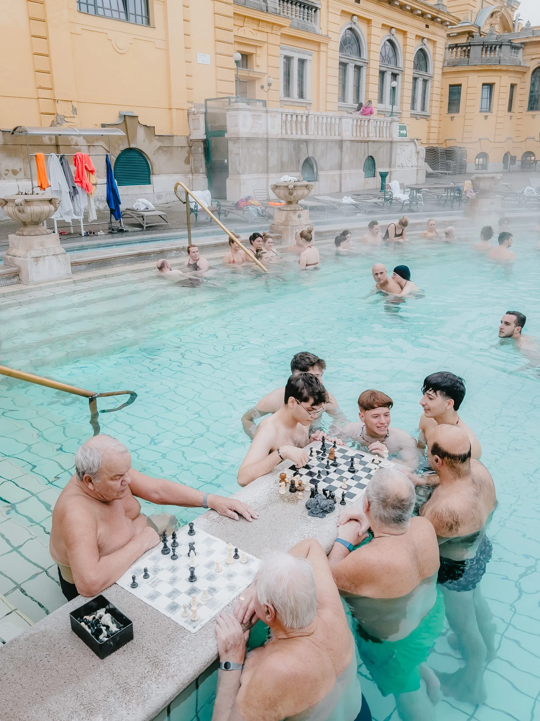 People playing chess by the poolside of a luxury hotel with classical architecture.