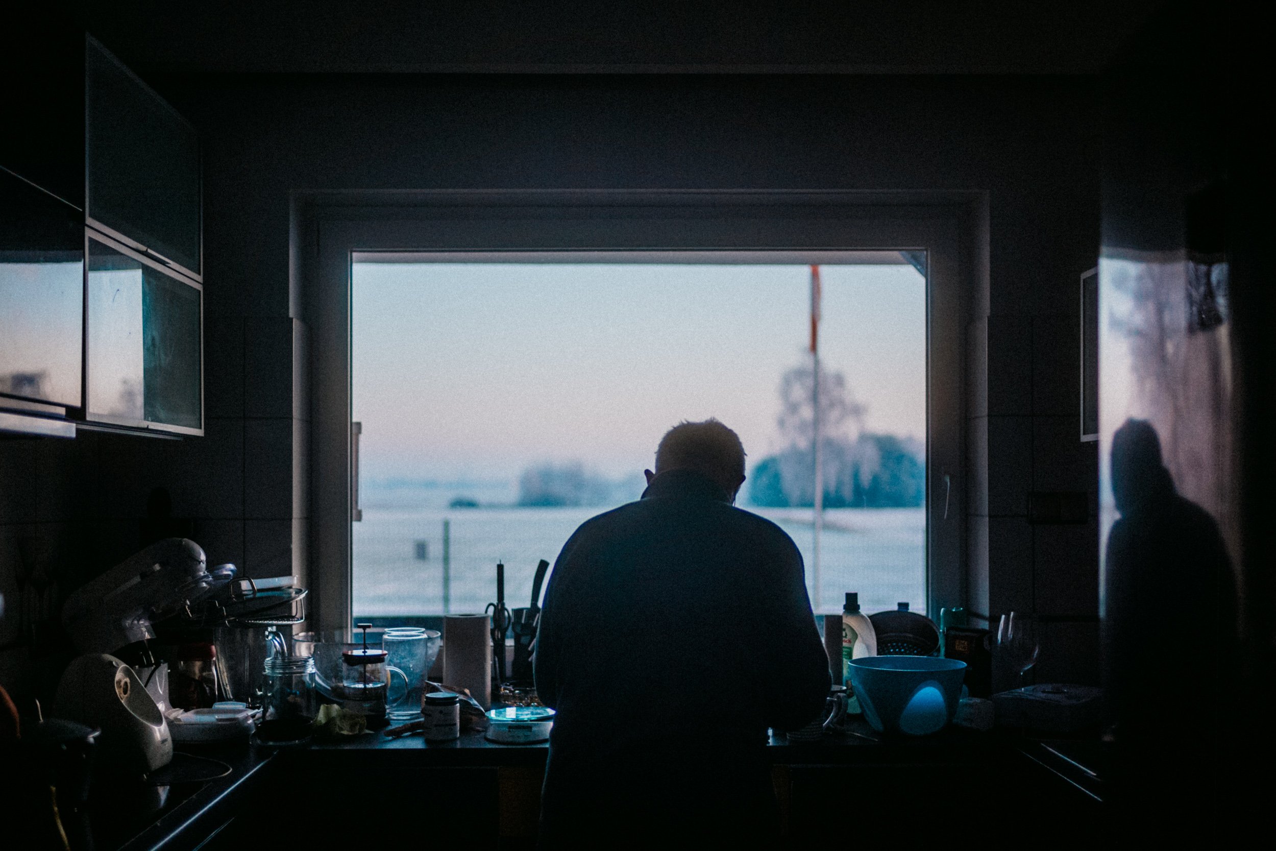 Inside a dark kitchen with a person facing a window, showing an outdoor scene with sky and distant structures, cluttered counter with various kitchen items.