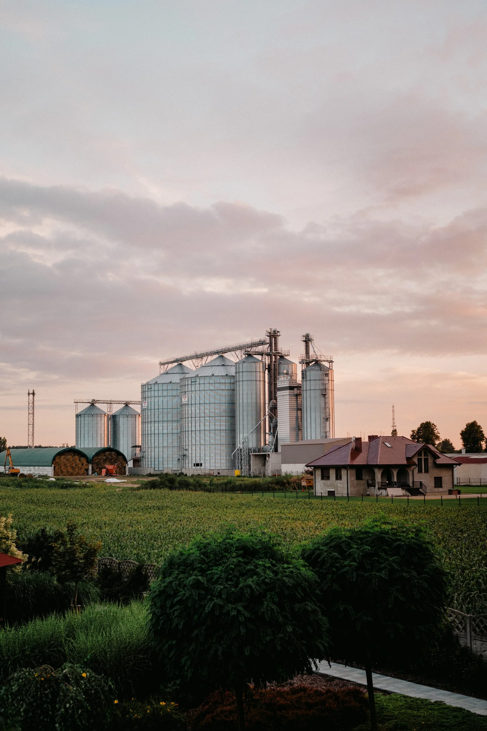 A farm scene at sunset with large silos and grain storage structures, a house with a red roof, and a lush green field in the foreground.