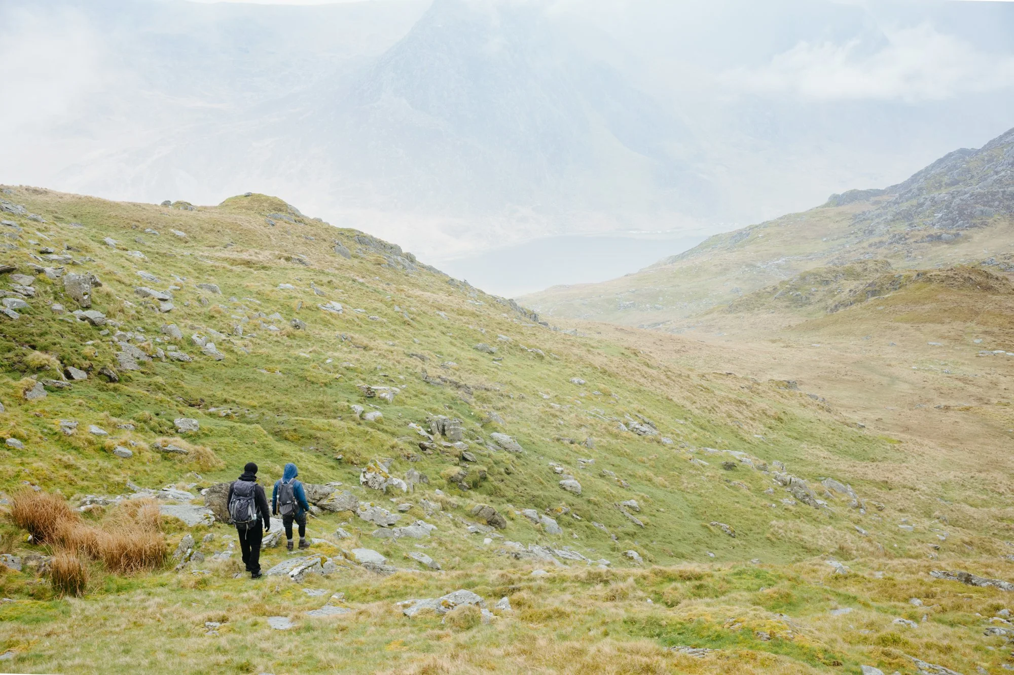 Two hikers with backpacks walking through a grassy mountainous landscape with clouds and distant mountains in the background.