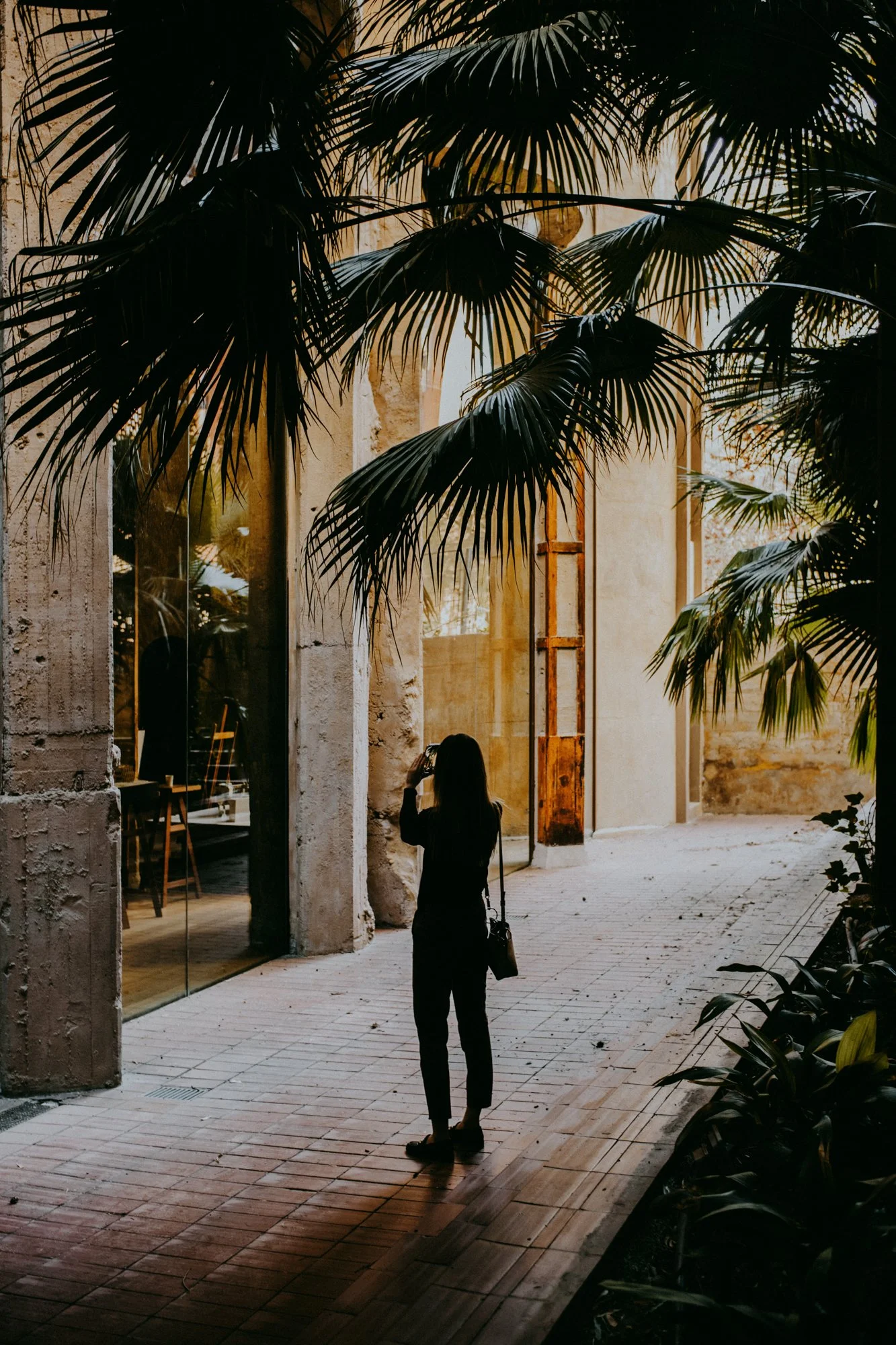 A woman standing outdoors taking a photo with a smartphone, surrounded by tropical plants and a rustic building with arches and large windows.