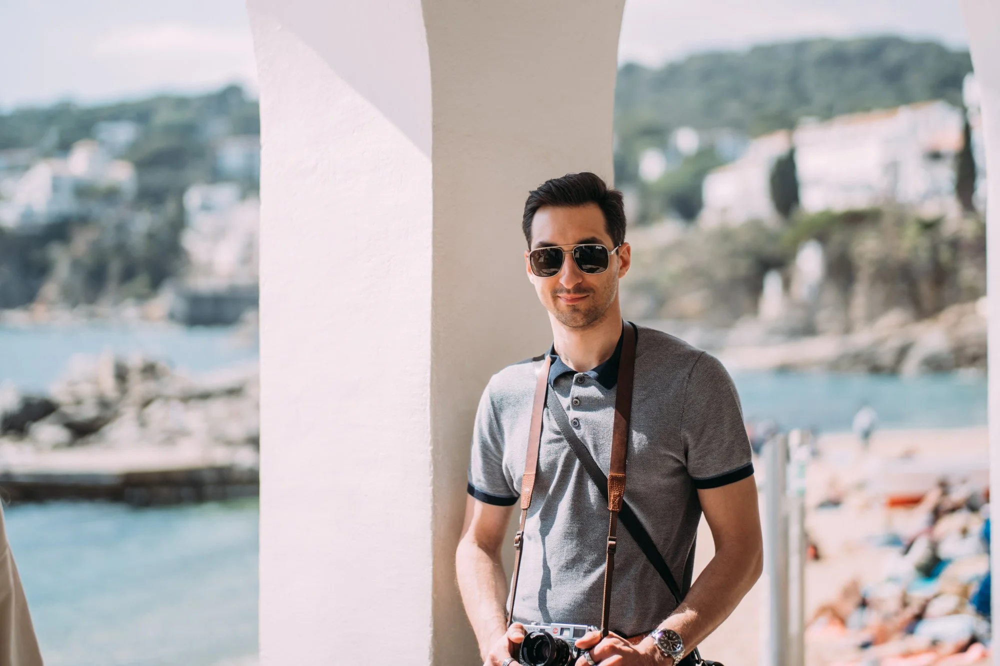 A young man wearing sunglasses and a gray polo shirt, holding a camera, standing outside near a white wall with a beach and hillside in the background.