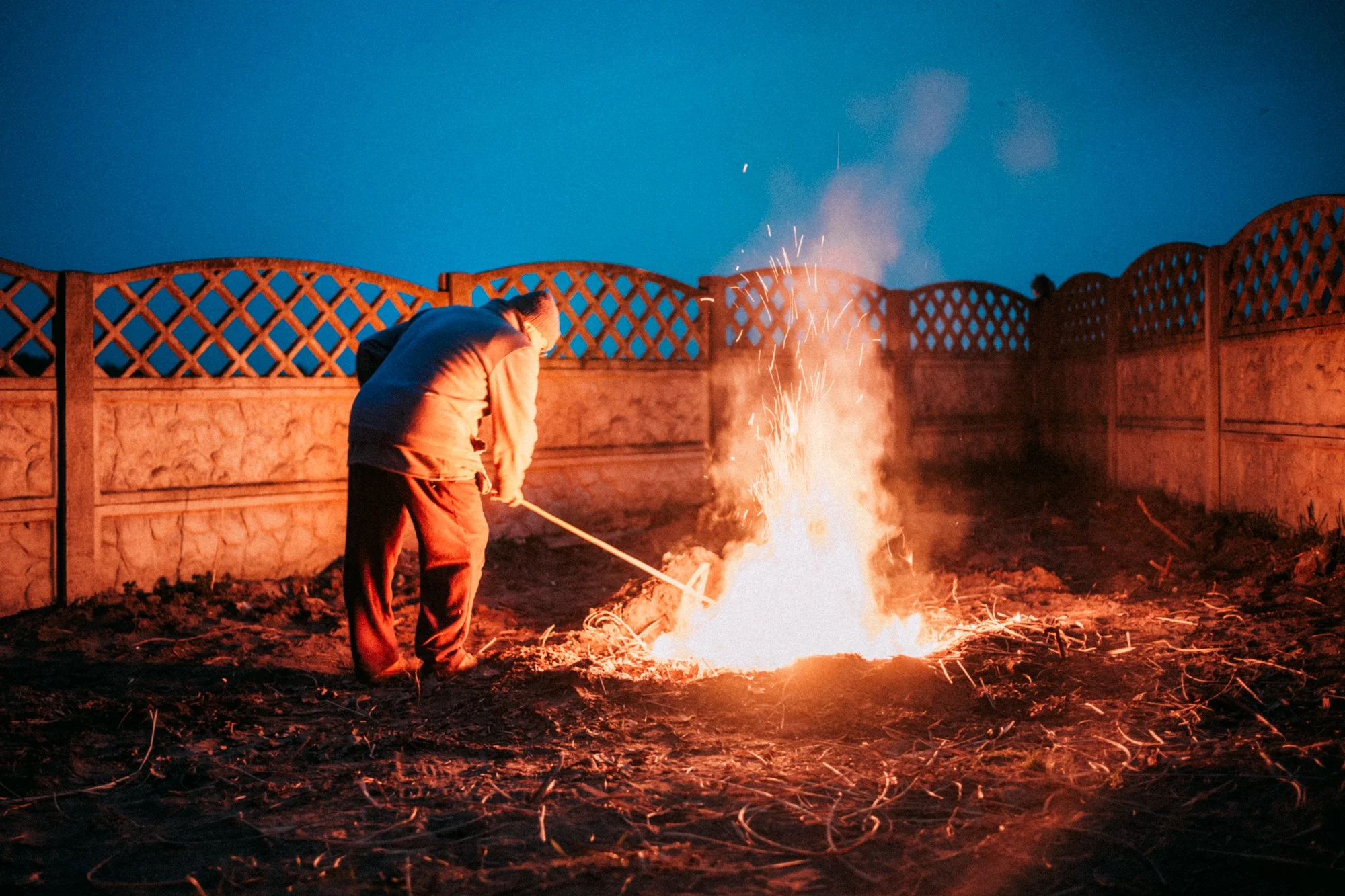Person wearing a hoodie and pants tending to a small controlled fire on dirt ground in a backyard at dusk, with a wooden lattice fence in the background.