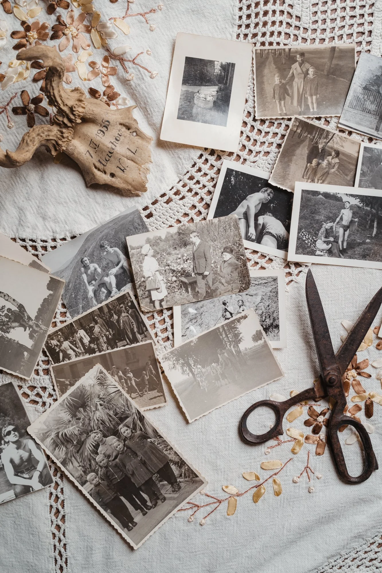 A collection of black and white vintage photographs scattered on a white crocheted tablecloth, alongside a pair of old scissors, a sea shell with writing on it, and small metallic leaf decorations.