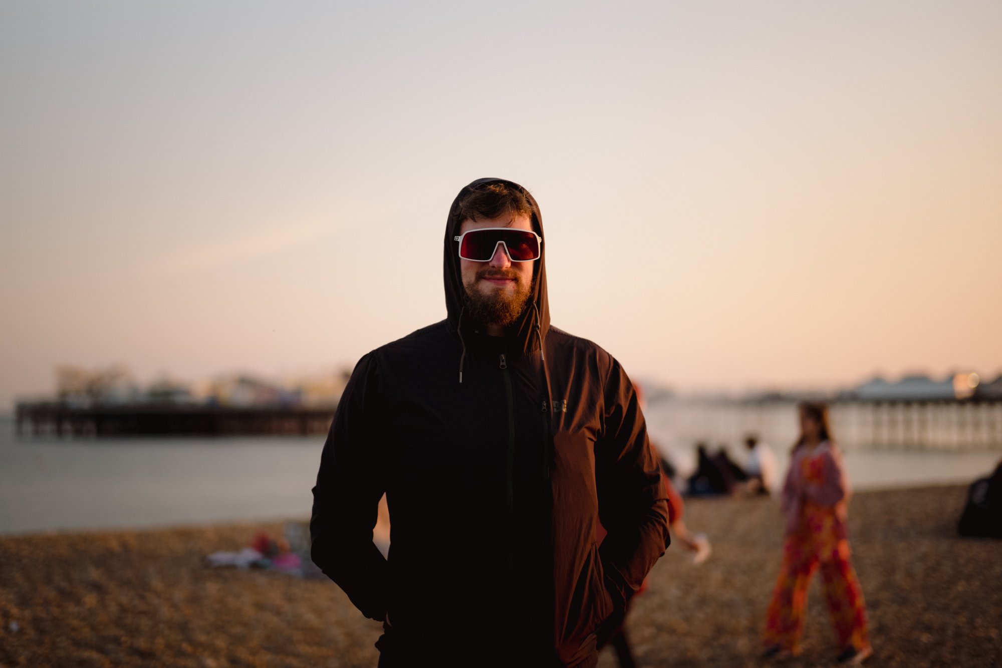 A man with a beard wearing a black hoodie and large sunglasses stands on a beach at sunset. In the background, there are blurry figures of people, including a woman in a floral dress, near the water.