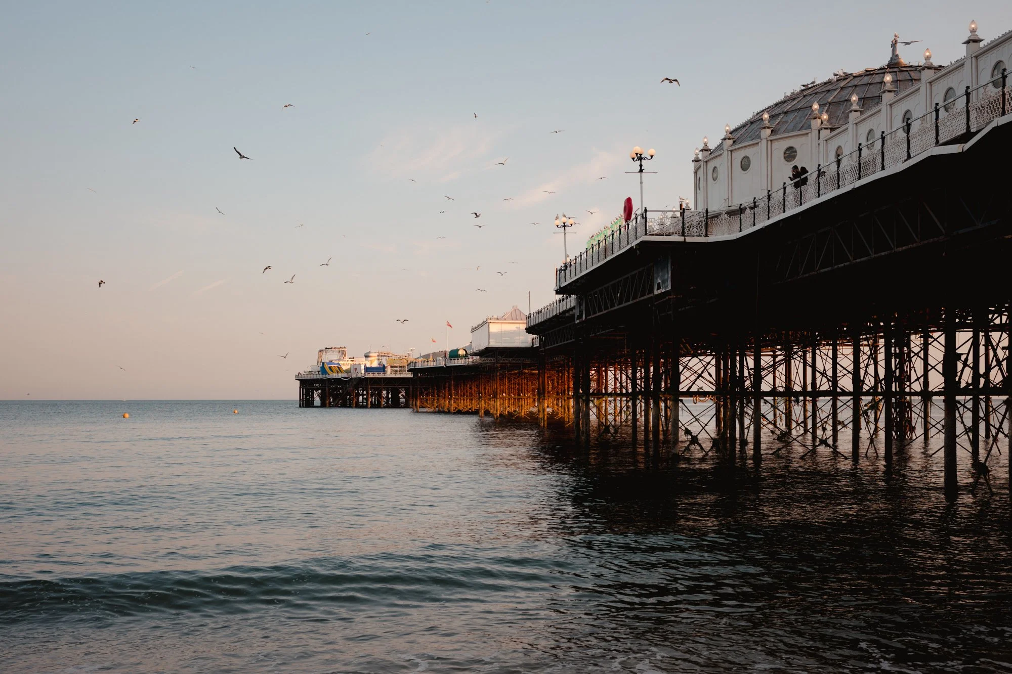 A pier extending into the ocean during sunset, with decorative white buildings and street lamps on top. Seagulls fly in the sky, and the water reflects the warm light of the sunset.