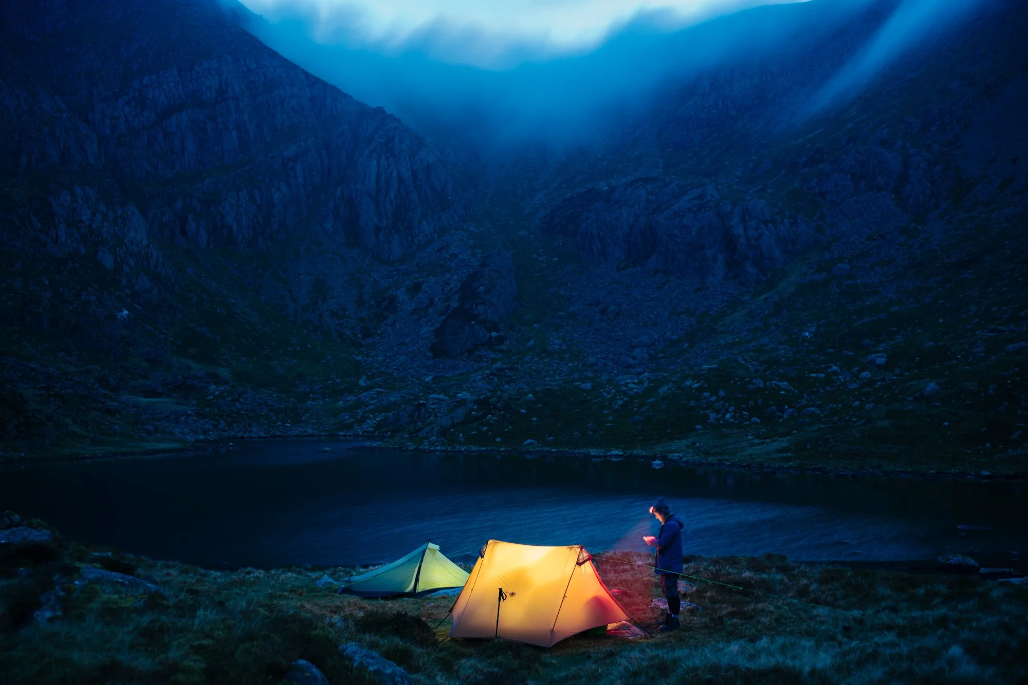 Nighttime scene of a person setting up a yellow tent near a lake in a mountainous area, with lakes and mountains in the background and a cloudy sky overhead.