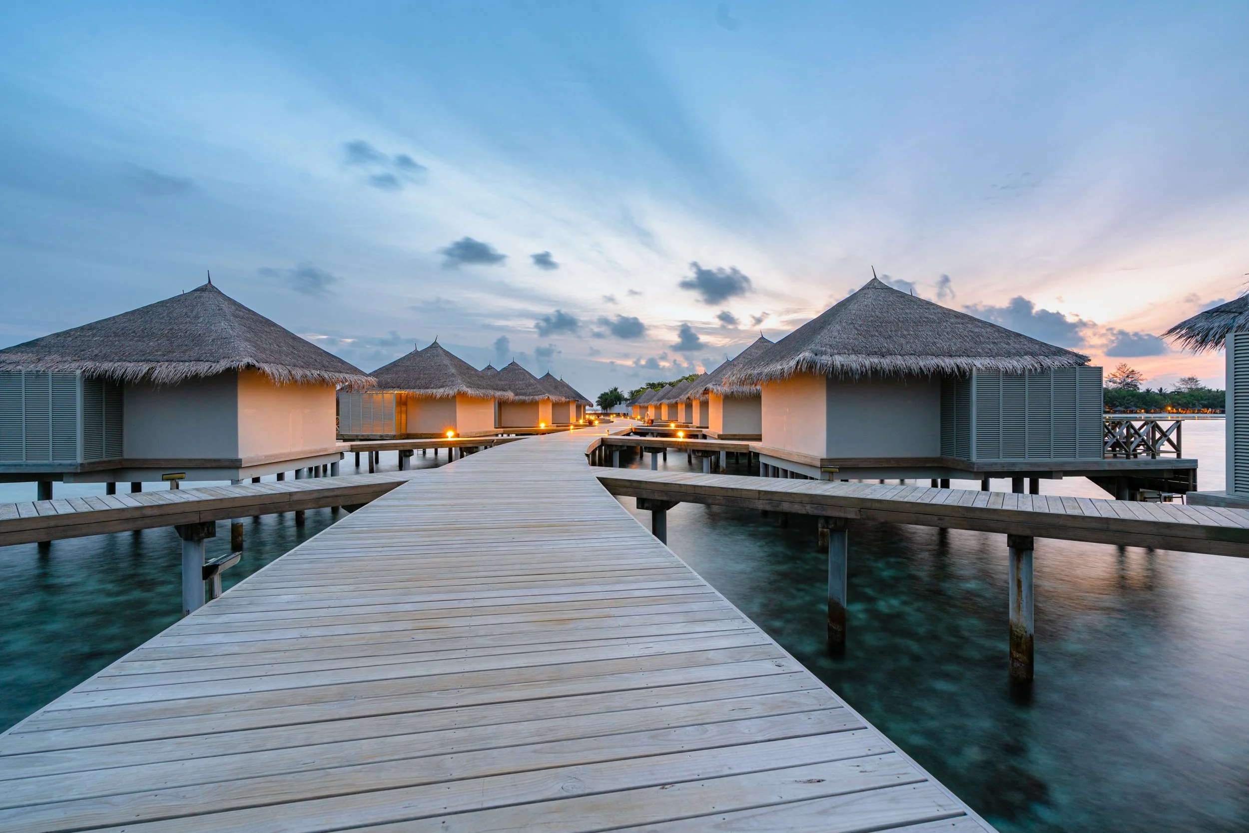 Overwater bungalows with thatched roofs connected by a wooden walkway, on a calm body of water during sunset with a partly cloudy sky.