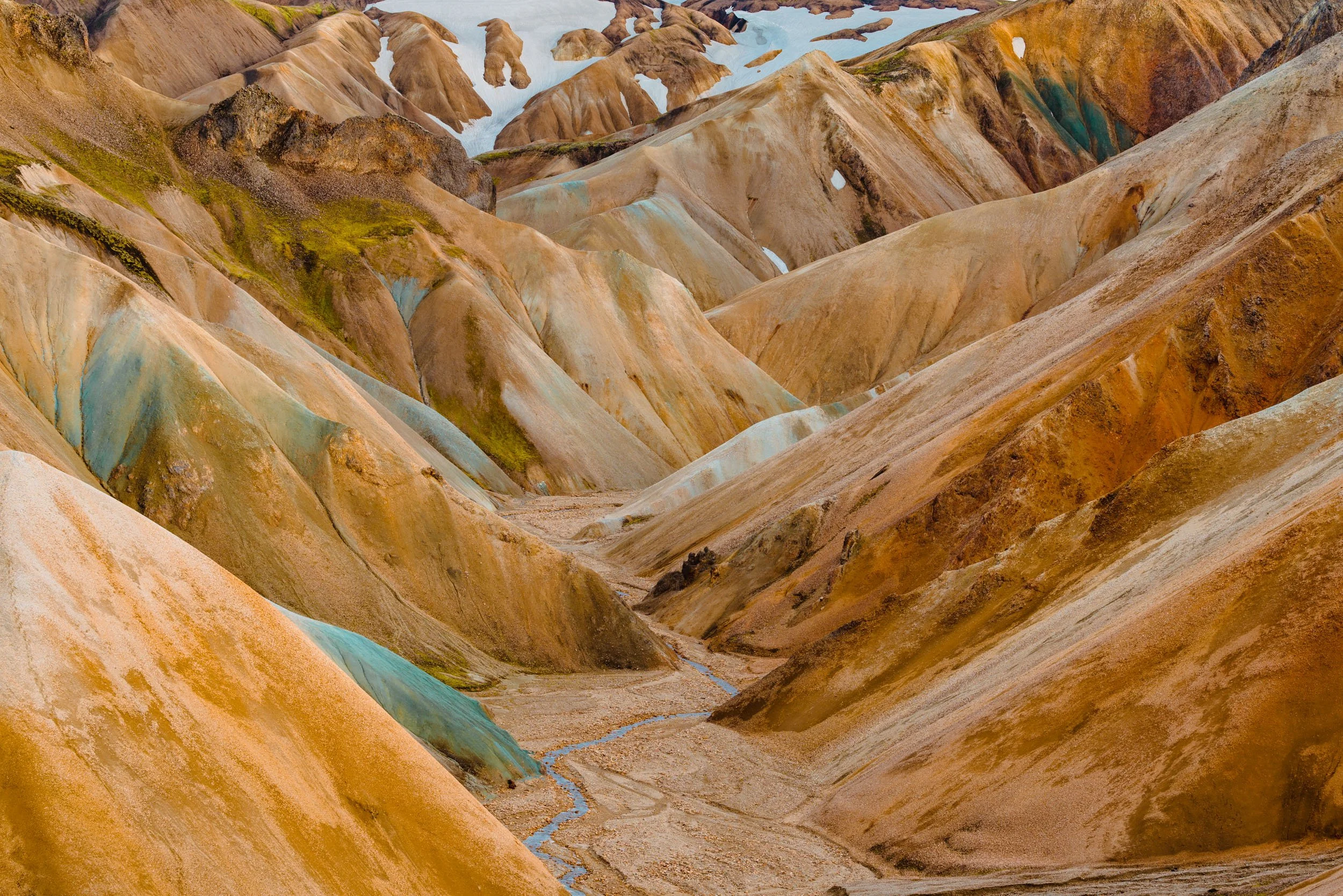 Colorful canyon with layered rock formations in shades of brown, orange, and blue, with a narrow stream flowing through the valley.