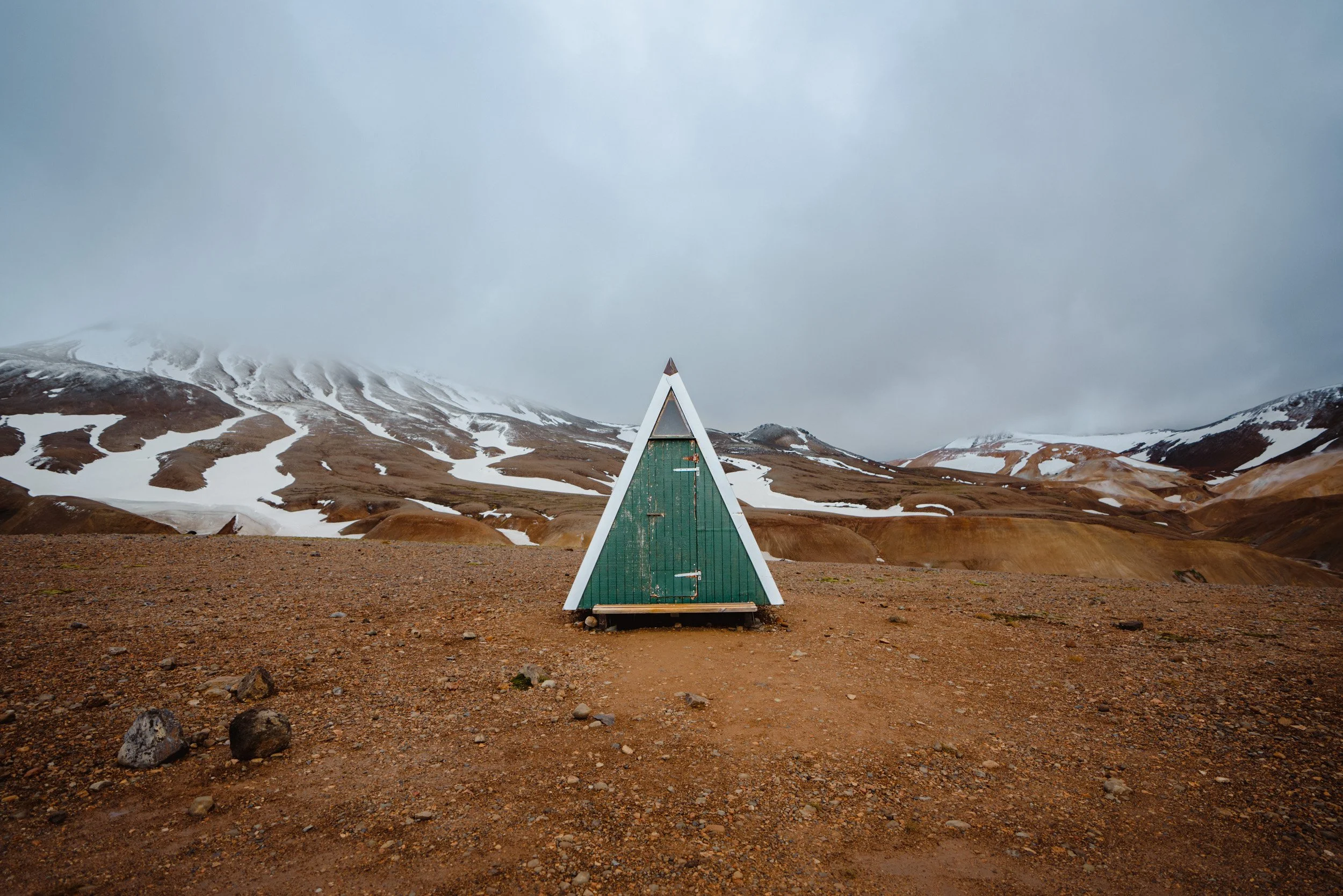 A small green triangular house on a flat, rocky terrain with a mountainous landscape, snow patches, and cloudy sky in the background.