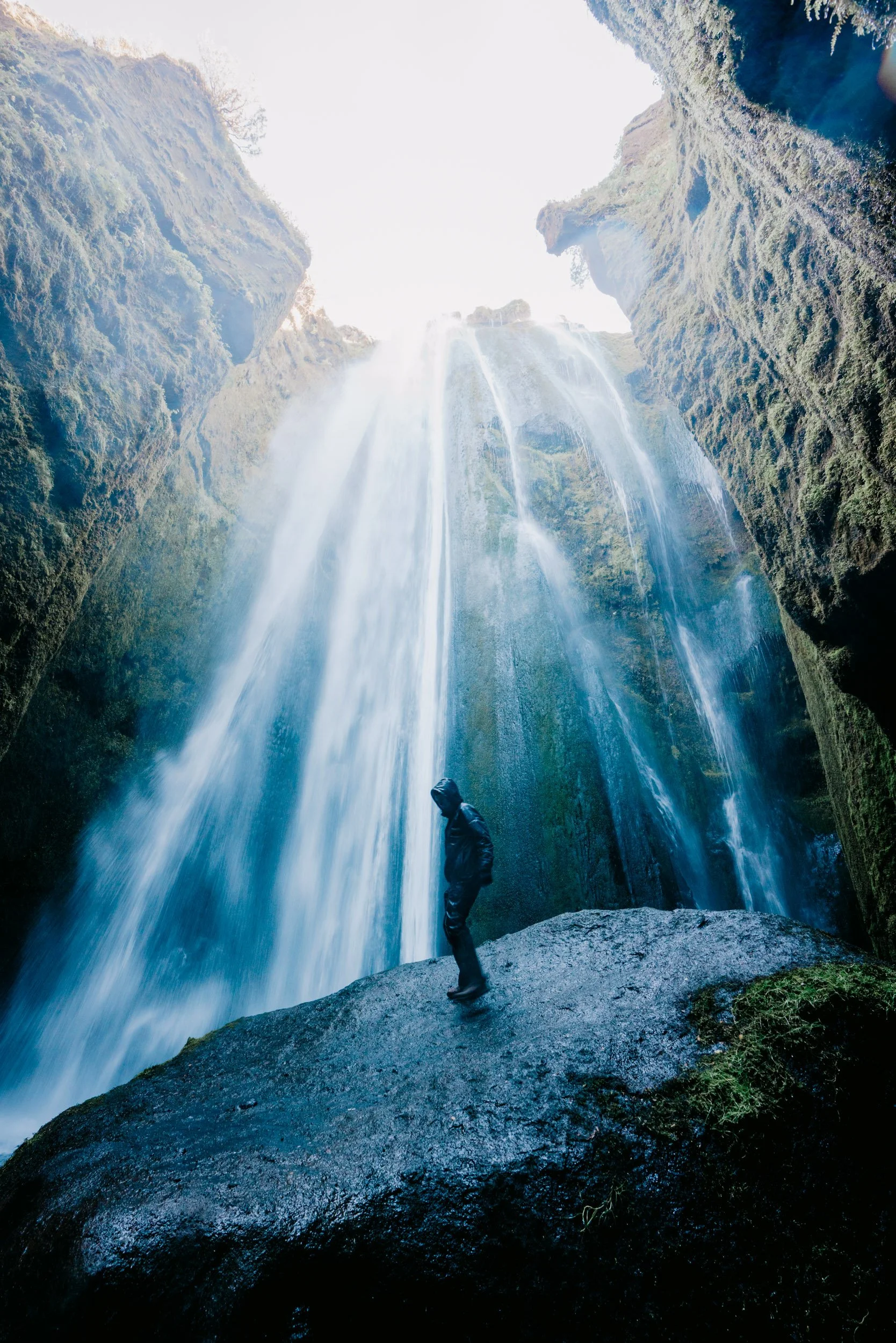 Person standing on a large rock at the base of a tall, cascading waterfall in a lush, green canyon with moss-covered rocks and steep cliffs.