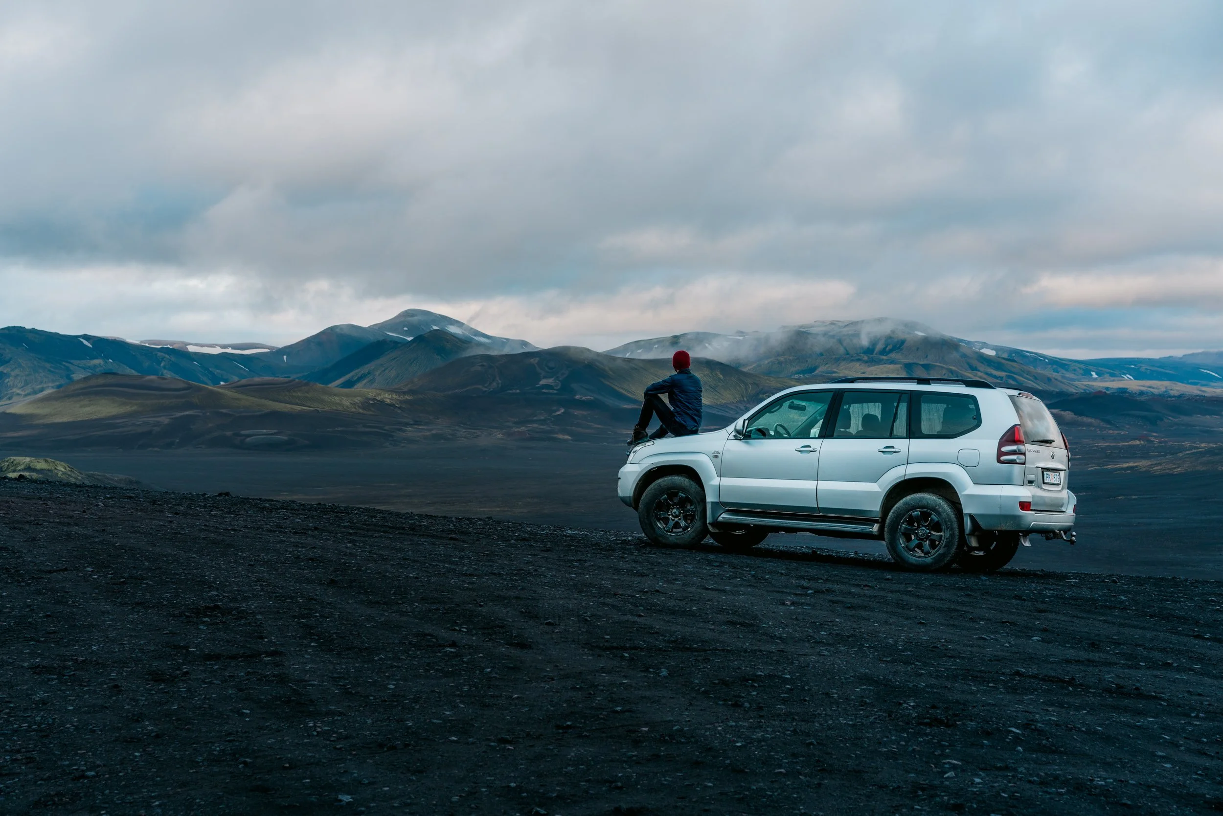A person sitting on the hood of a white SUV parked on a dark gravel surface, overlooking mountainous terrain under a cloudy sky. The person wears a red beanie and a dark jacket, facing away from the camera.