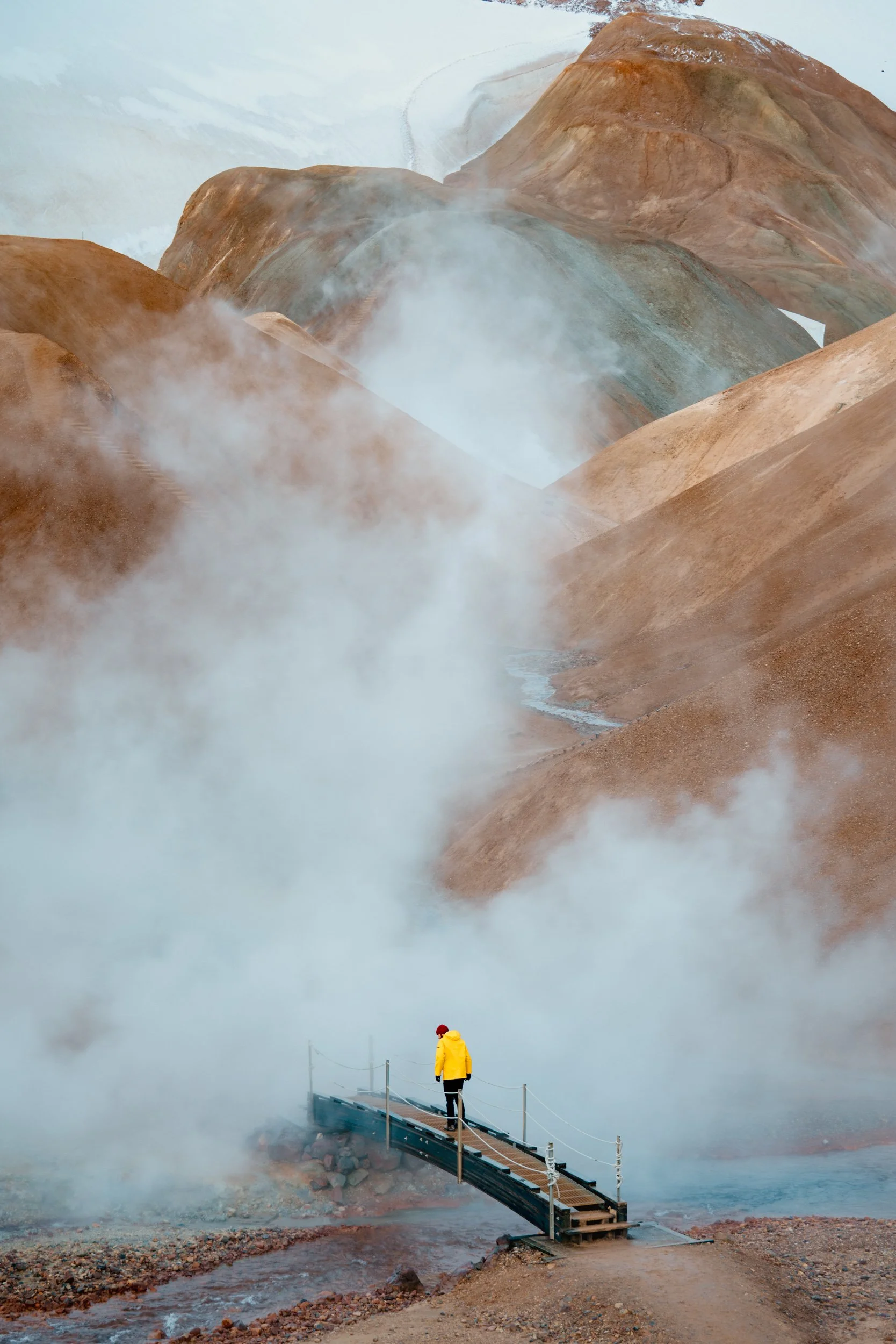 A person in a yellow jacket and red hat walking on a small bridge over steaming geothermal vents, with colorful rocky hills in the background.