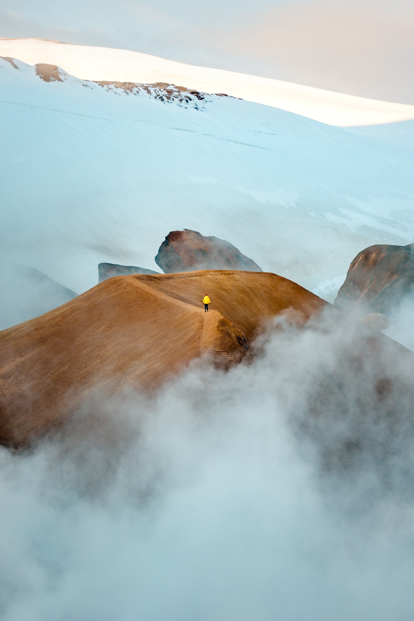 A person in a yellow jacket walking on a narrow brown trail through mountainous terrain with snow-capped peaks in the background