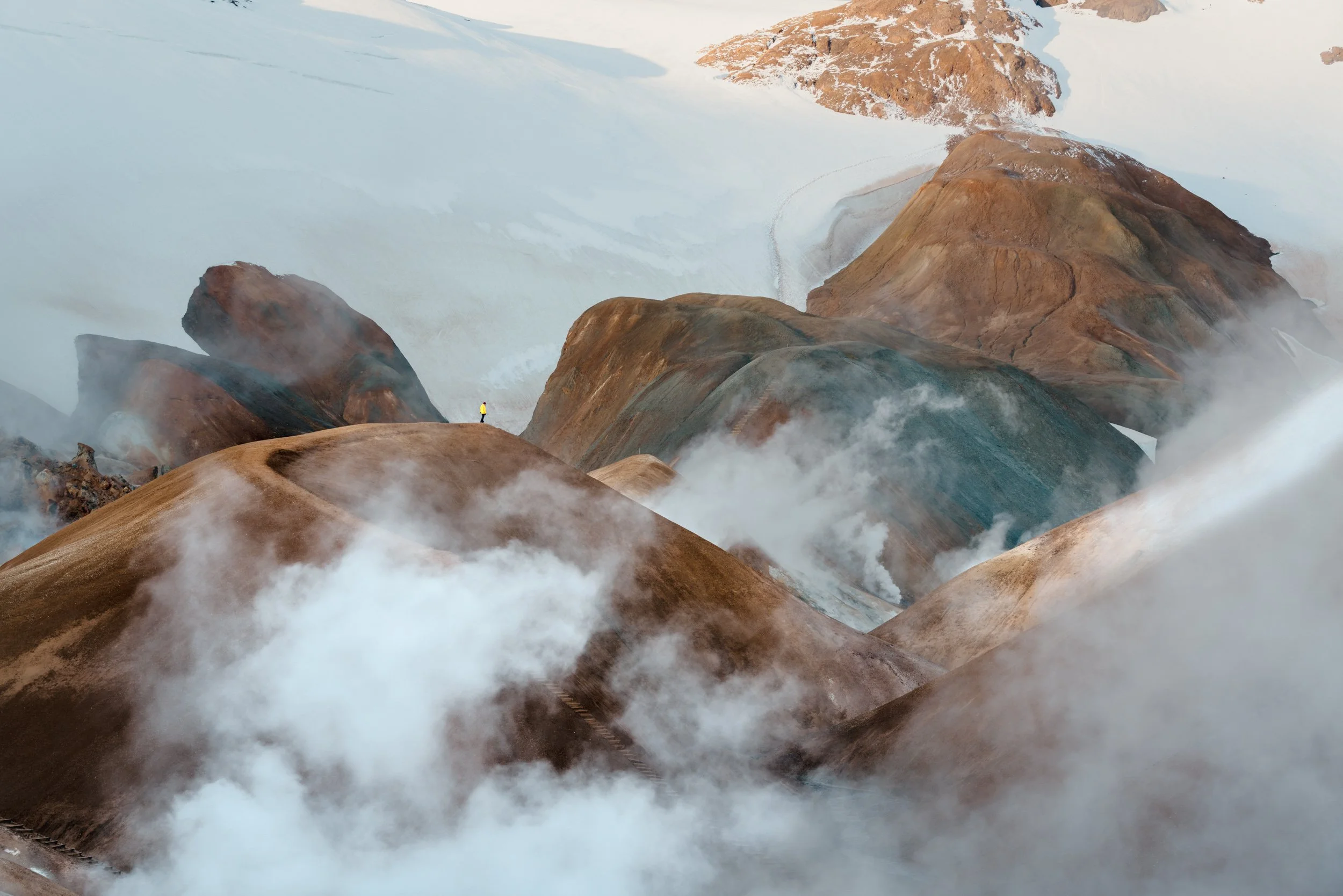 A mountain landscape with brown and reddish rocky formations, some snow patches, and clouds or mist surrounding the mountains.