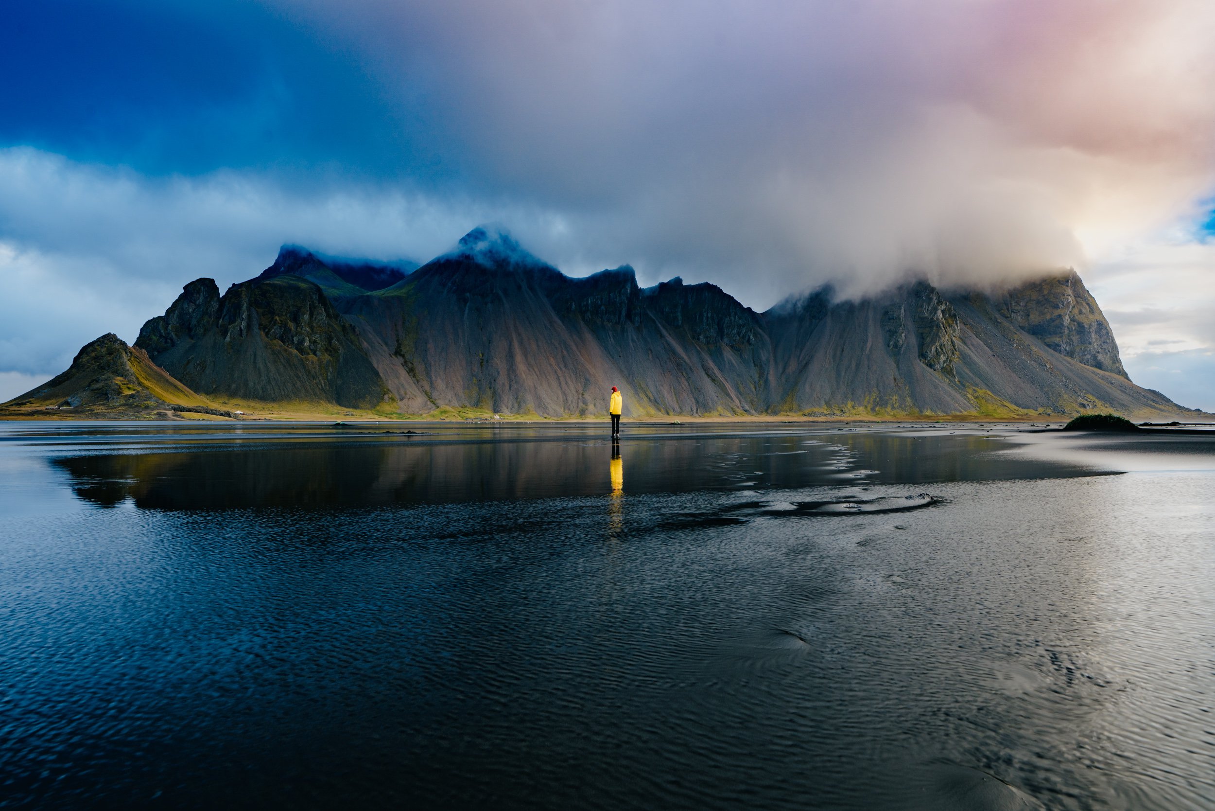 A person wearing a yellow jacket and red hat standing at the edge of a body of water with mountain peaks and overcast sky in the background.