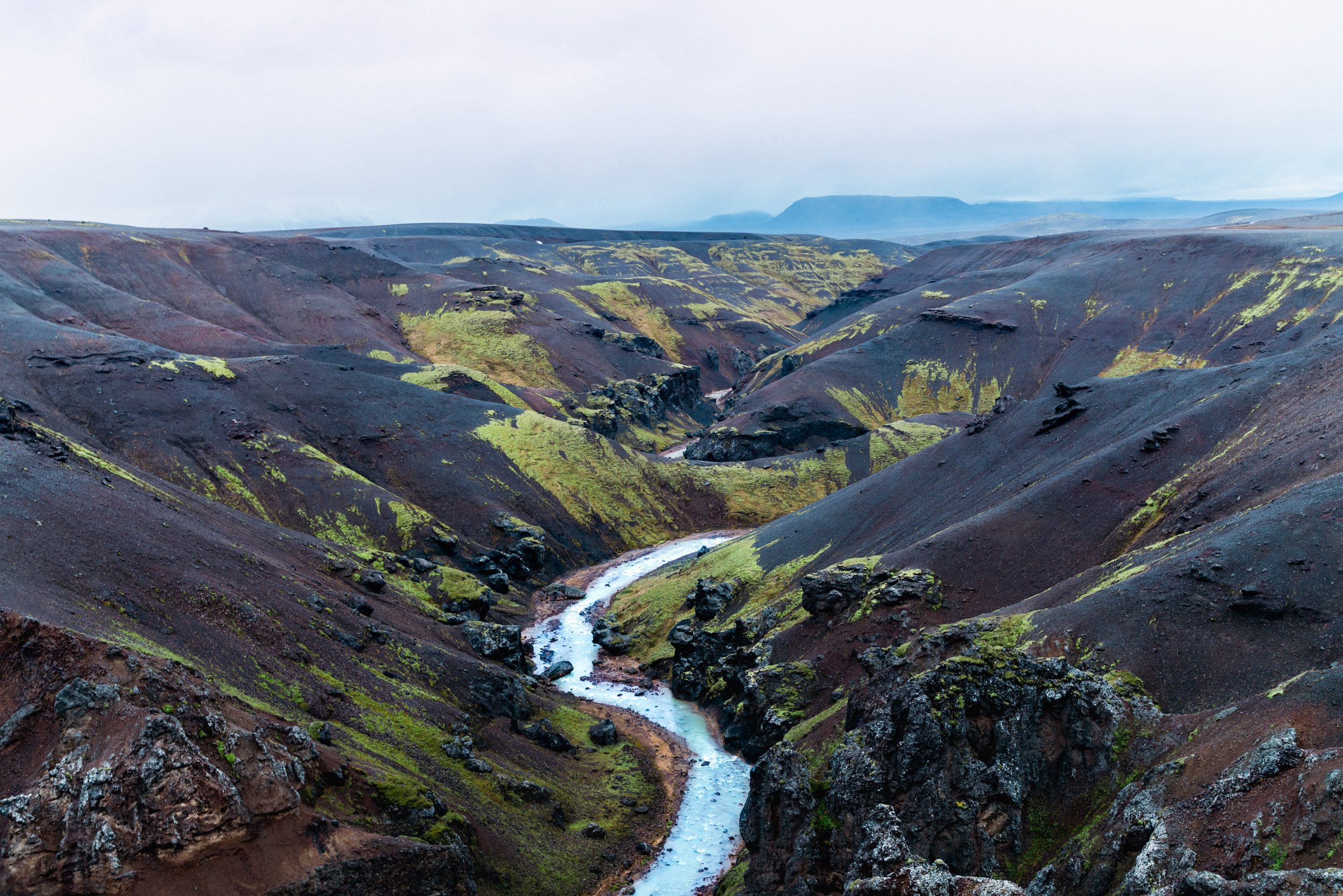 A rugged canyon with dark volcanic rock and patches of green moss, featuring a winding stream flowing through the center under a cloudy sky.
