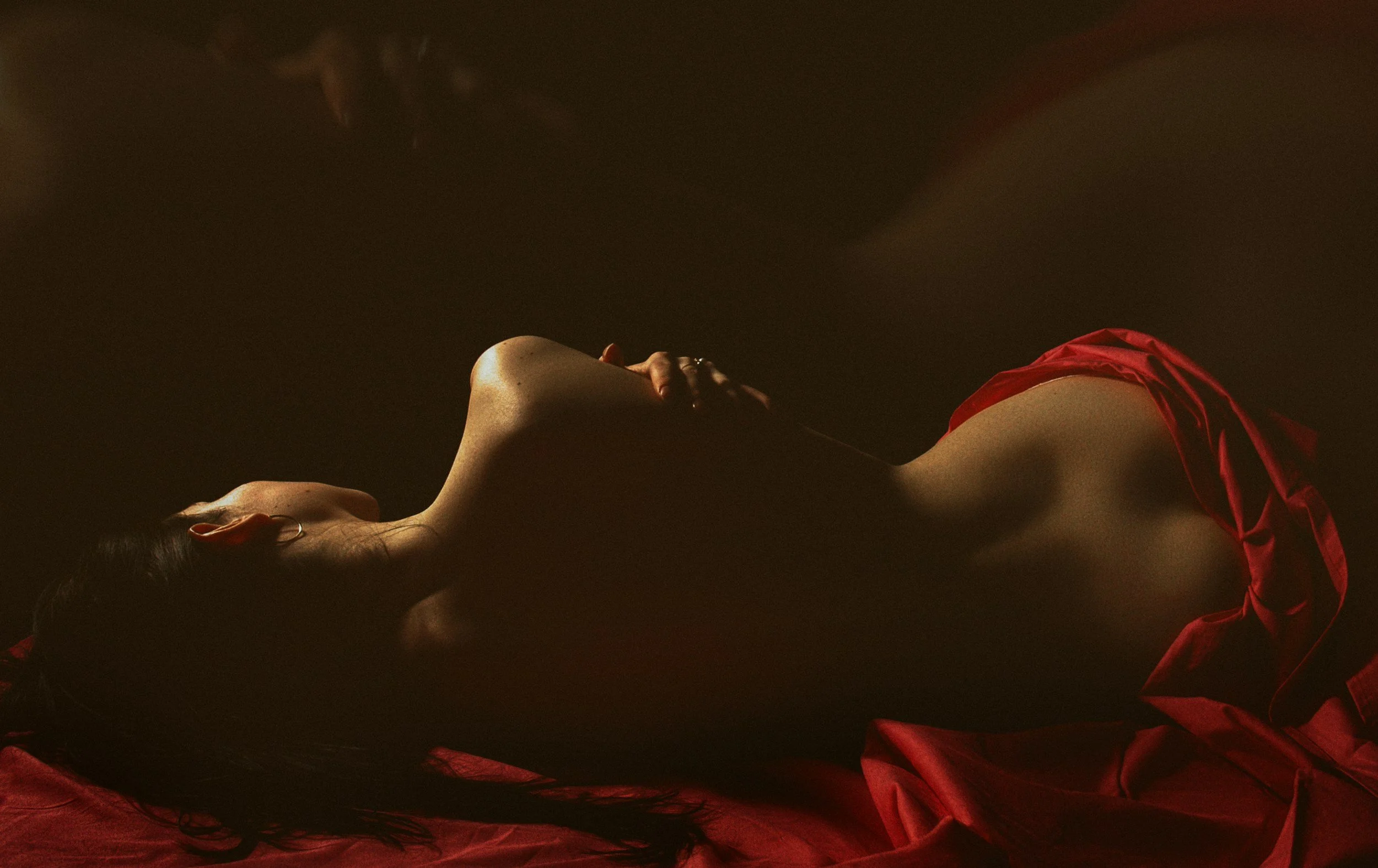 A woman lying on her side in dark lighting, wearing a red satin garment, with long dark hair, and her hand resting on her chest.