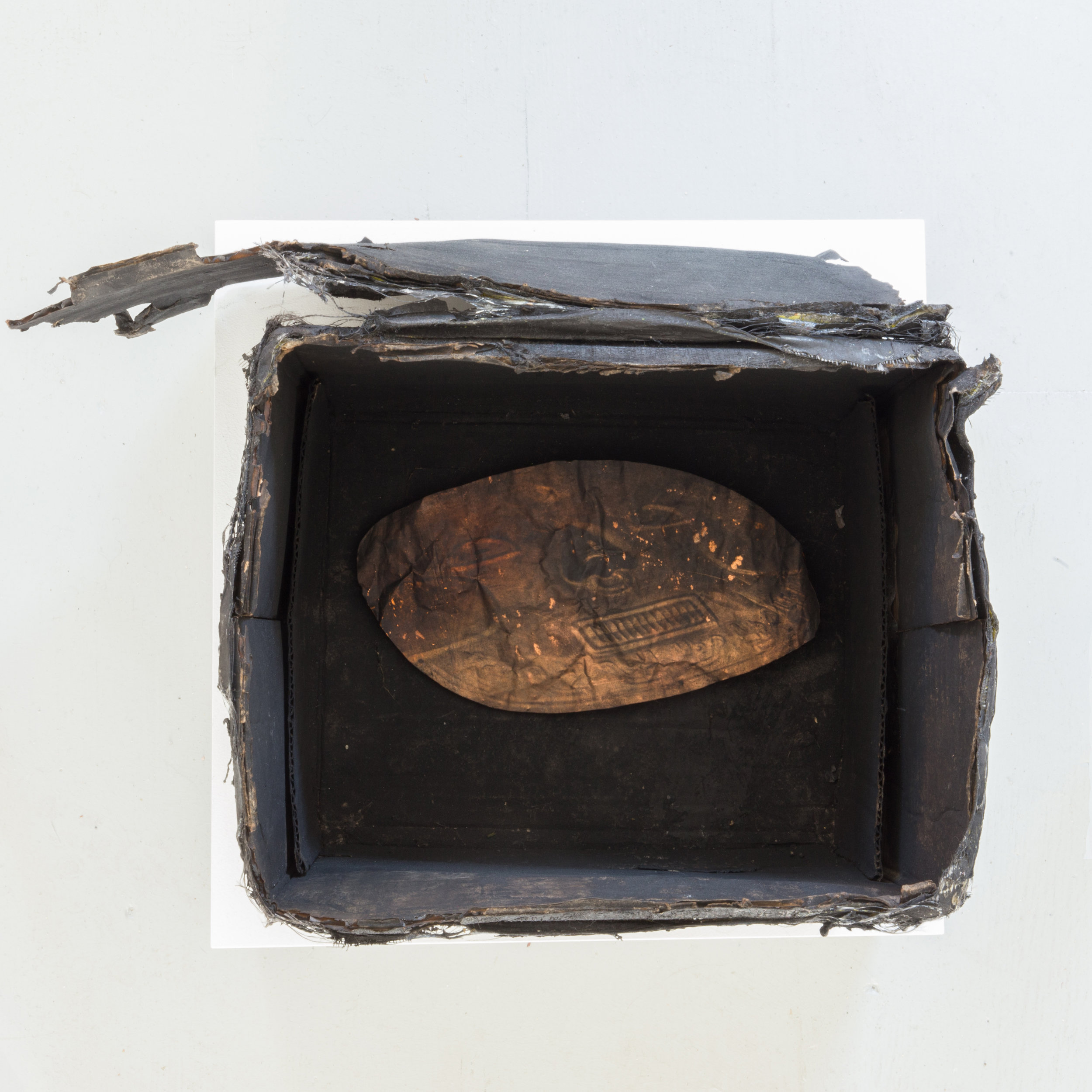 Harold Mendez. "Untitled (Death Mask)," 2015. Burned cardboard box, soot, toner, oxidized copper reproduction of pre-Columbian death mask from the Museo del Oro (Bogota, Colombia). 14" x 20" x 14"