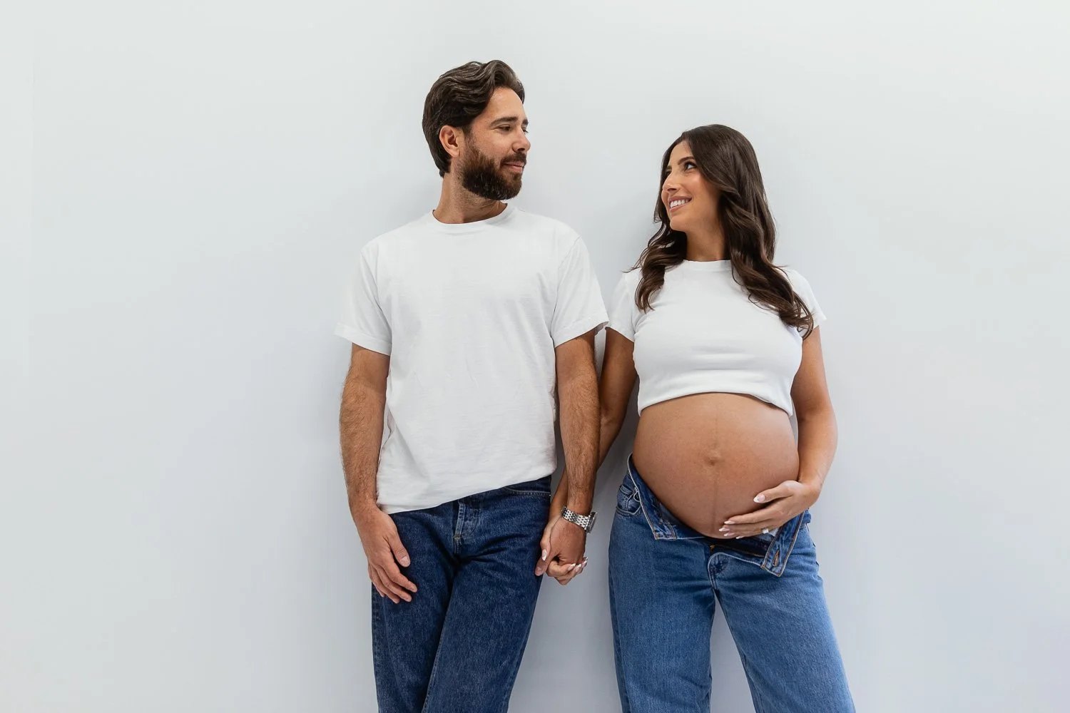 A pregnant woman holding her belly and smiling at a man, they are standing against a plain white wall, holding hands, both dressed in white T-shirts and jeans. maternity photoshoot.
