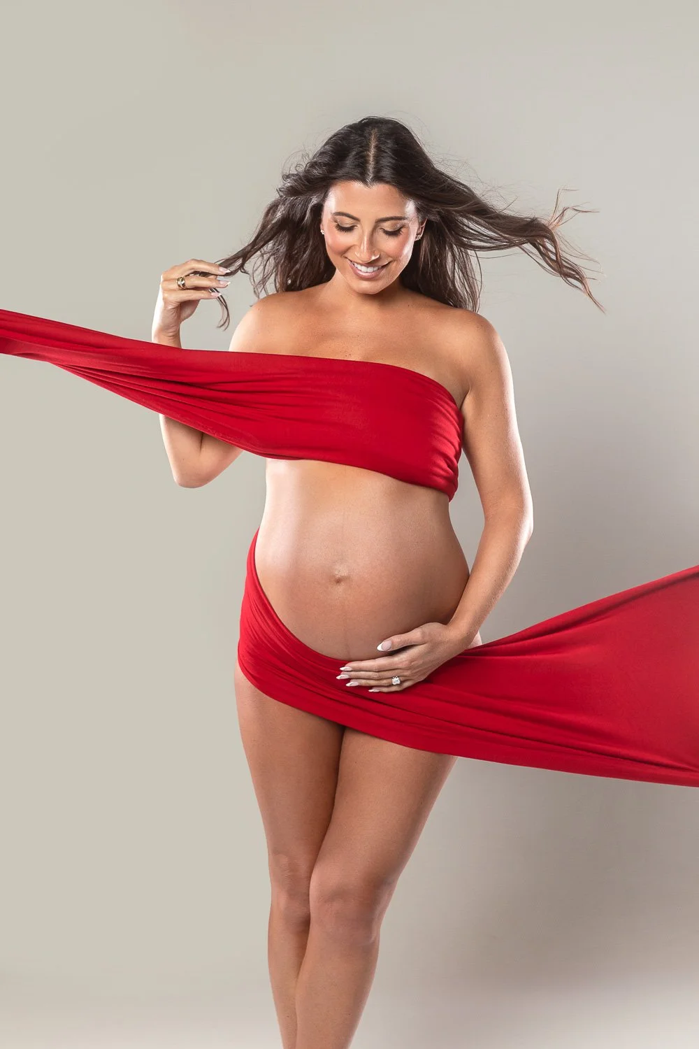 Pregnant woman with long, flowing brown hair smiling, wrapped in red cloth, posing against a plain light gray background. maternity photoshoot.