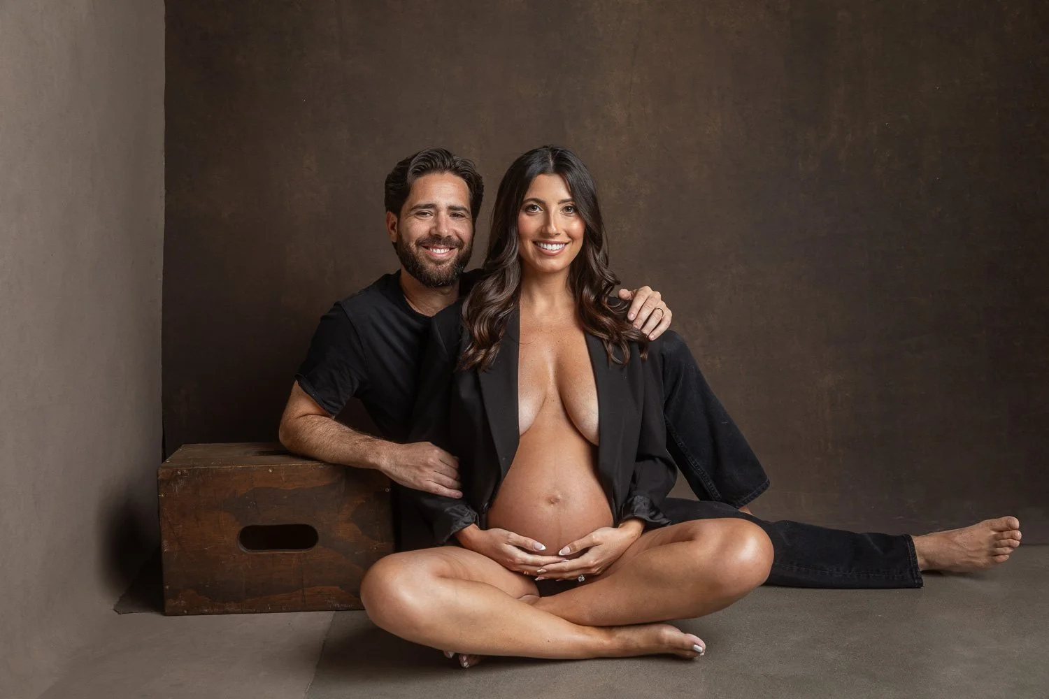 A pregnant woman with long dark hair and a man with dark hair and beard sitting on the floor, smiling at the camera, with the woman's hand on her belly and the man's arm around her shoulder, in front of a brown backdrop. maternity photoshoot.