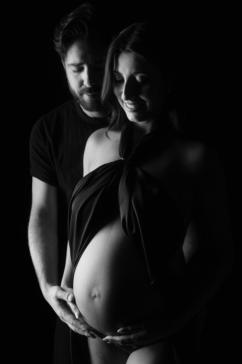 A pregnant woman with a man standing behind her, gently holding her belly. They are both smiling softly. The image is in black and white, with a dark background. maternity photoshoot.
