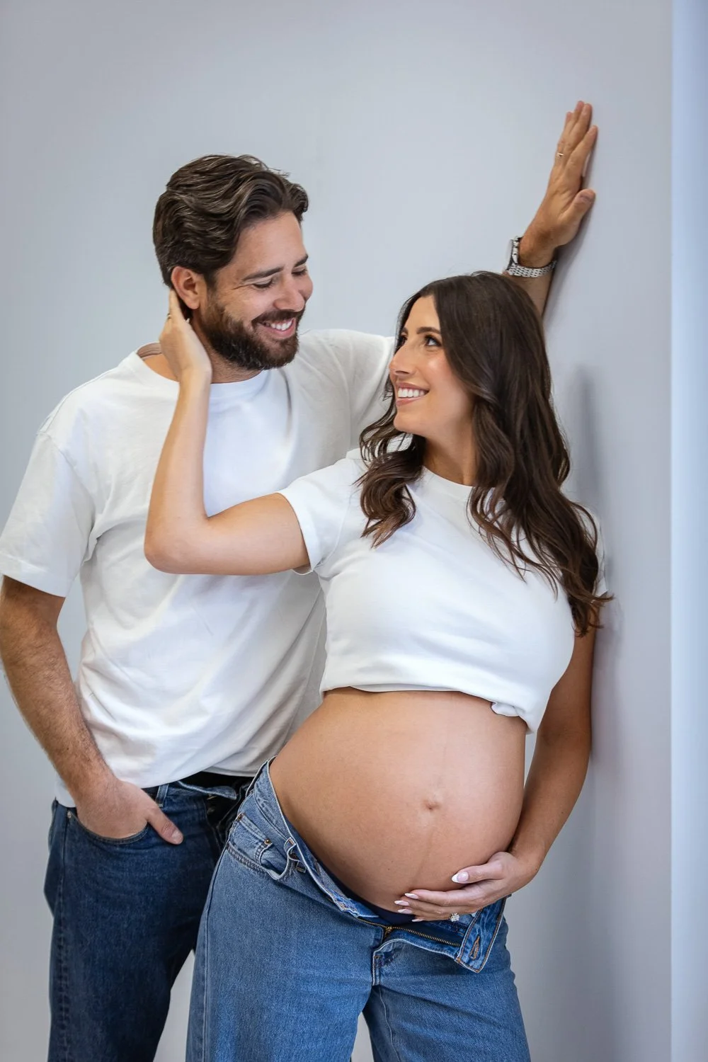 A happy couple, with a pregnant woman and a man, sharing a moment together against a plain wall. The woman is wearing a white T-shirt lifted to reveal her pregnant belly, and the man has his hand on her belly and is leaning against the wall with his 